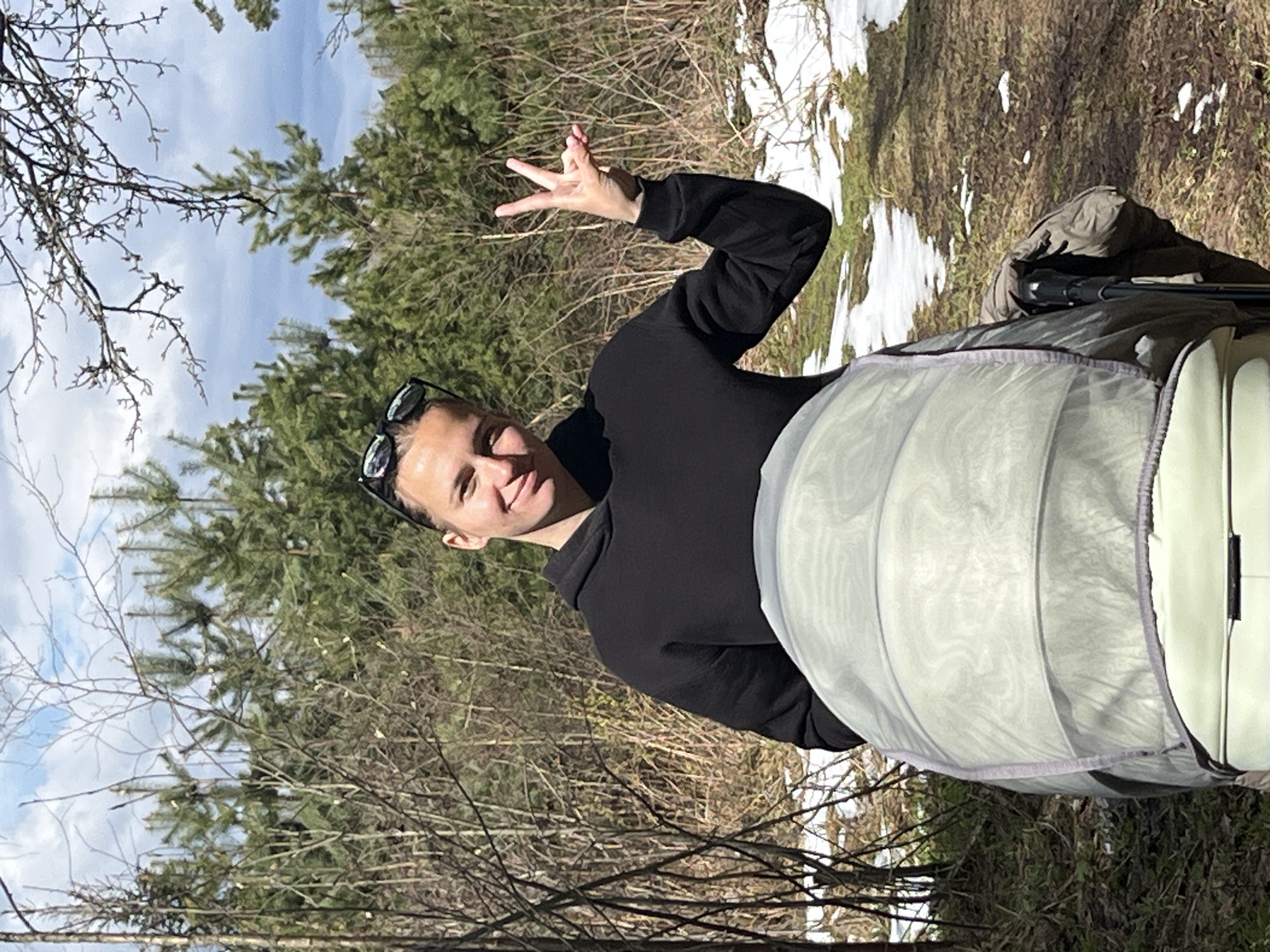 A woman is standing outside in a wooded area near Раздолье, Russia. She is wearing a black hoodie and sunglasses on her head, and is making a peace sign with her right hand. A baby stroller is next to her. The background includes trees, bushes, and some snow on the ground. The sky is visible with some clouds.