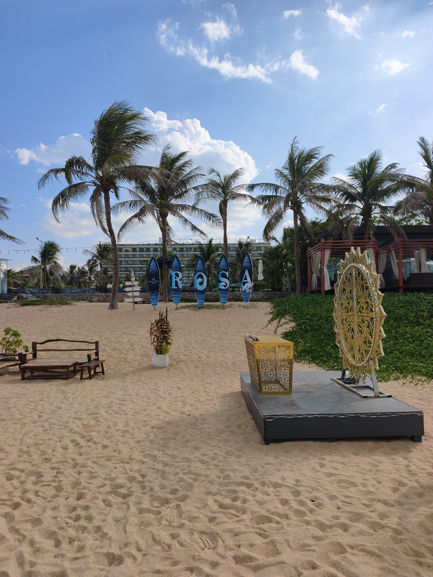 The image depicts a sandy beach in Tuy Hoa, Vietnam, with several surfboards standing upright and spelling out "ROSA". In the foreground, a wooden bench and table set rests on the sand. Behind the surfboards, a modern building with multiple floors is visible, surrounded by palm trees. To the right, a decorative golden wheel-like structure is placed on a platform. Lush green vegetation covers a slope on the right side of the scene. The sky is blue with scattered white clouds, and a faint airplane contrail is visible in the distance.
