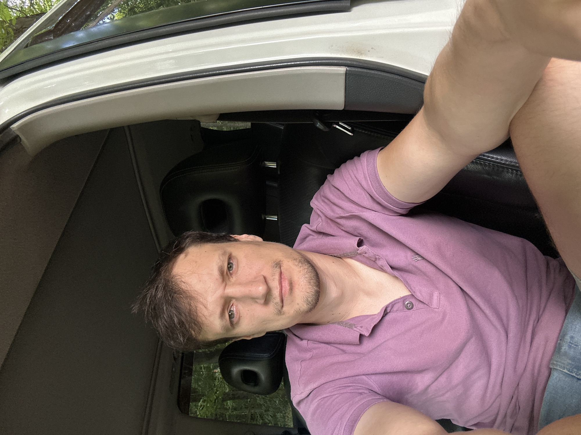 A man with short brown hair and stubble is seated in the driver's seat of a white SUV. He is wearing a light purple polo shirt and blue denim shorts. The interior of the SUV is visible, including the black leather seats and part of the dashboard. Outside the vehicle, trees and foliage suggest a natural, possibly wooded, environment. The image is a selfie, taken from the man's perspective. The location of Shatava, Ukraine, is not discernible from the image.
