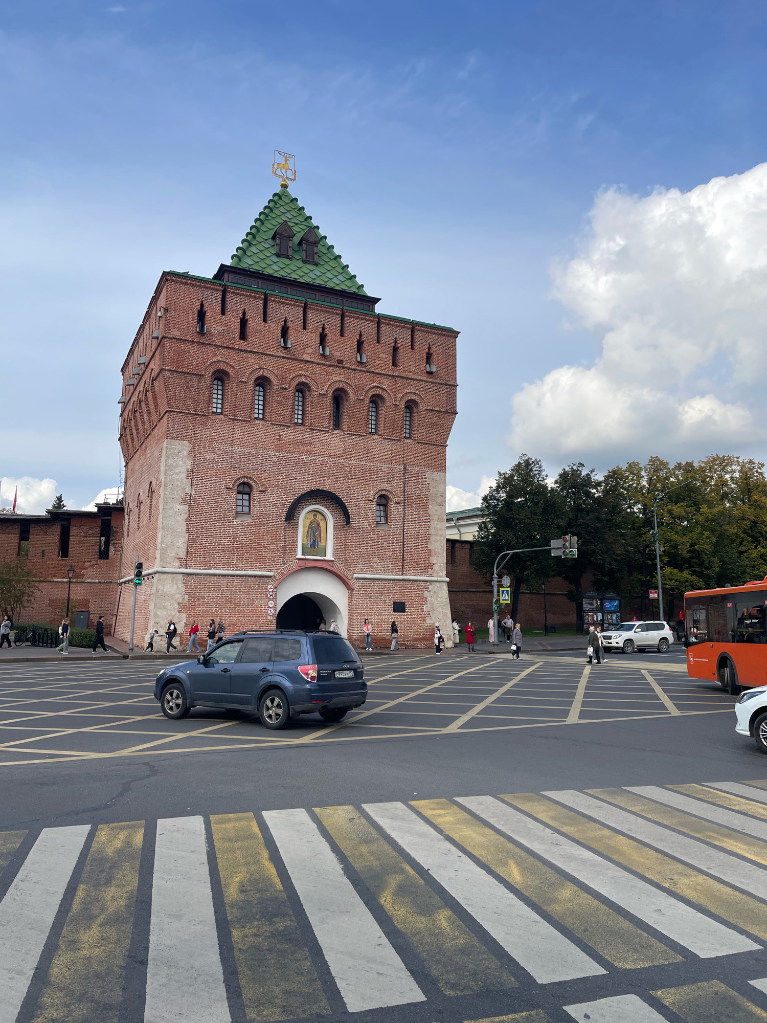 The image shows the Ivanovo Tower, a prominent landmark in the Nizhny Novgorod Kremlin, Russia. The tower is constructed of red brick and has a green, pyramidal roof topped with a golden emblem of a horseman. In the foreground, a blue Subaru Forester is stopped at a pedestrian crossing, with yellow and white stripes painted on the asphalt. Other vehicles, including an orange bus and a white SUV, are visible in the background, along with several pedestrians crossing the street. The sky is partly cloudy, with a mix of blue sky and white cumulus clouds. Trees with green foliage are visible behind the tower and to the right of the image. The overall scene depicts a busy urban intersection in front of a historic fortification.