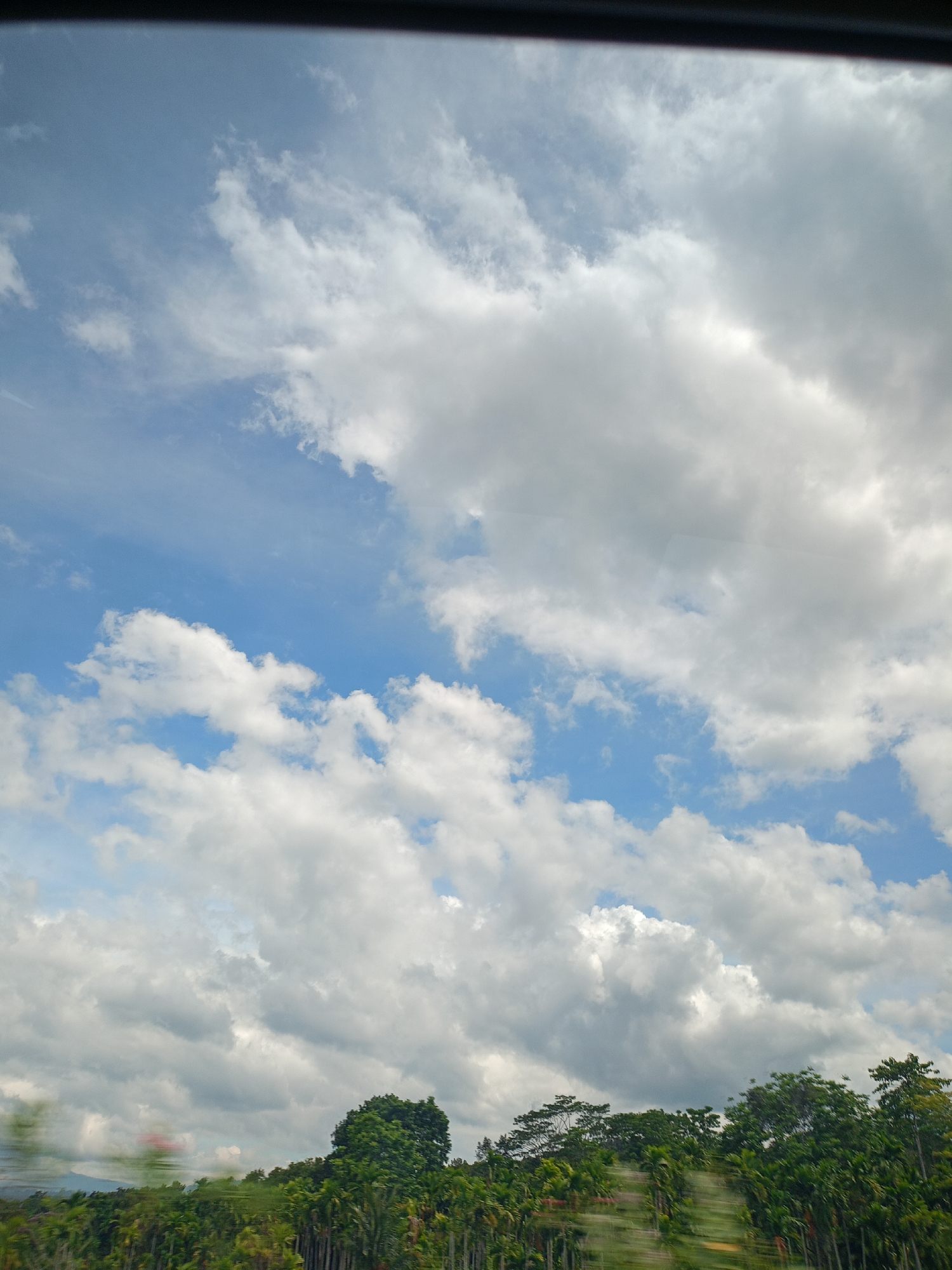 The photograph presents a view of the sky and treetops, likely taken from a moving vehicle in Grong-Grong, Indonesia. The sky is predominantly blue with numerous white cumulus clouds scattered throughout. The cloud cover varies in density, with some areas showing clear blue sky and others obscured by thick cloud formations. The bottom portion of the image shows a line of green trees, blurred, suggesting motion of the vehicle. The blurred red and green shapes in the lower left may be other moving vehicles. A portion of the vehicle's frame is visible at the top edge of the photo. The scene conveys a sense of movement, and a bright, partly cloudy day.