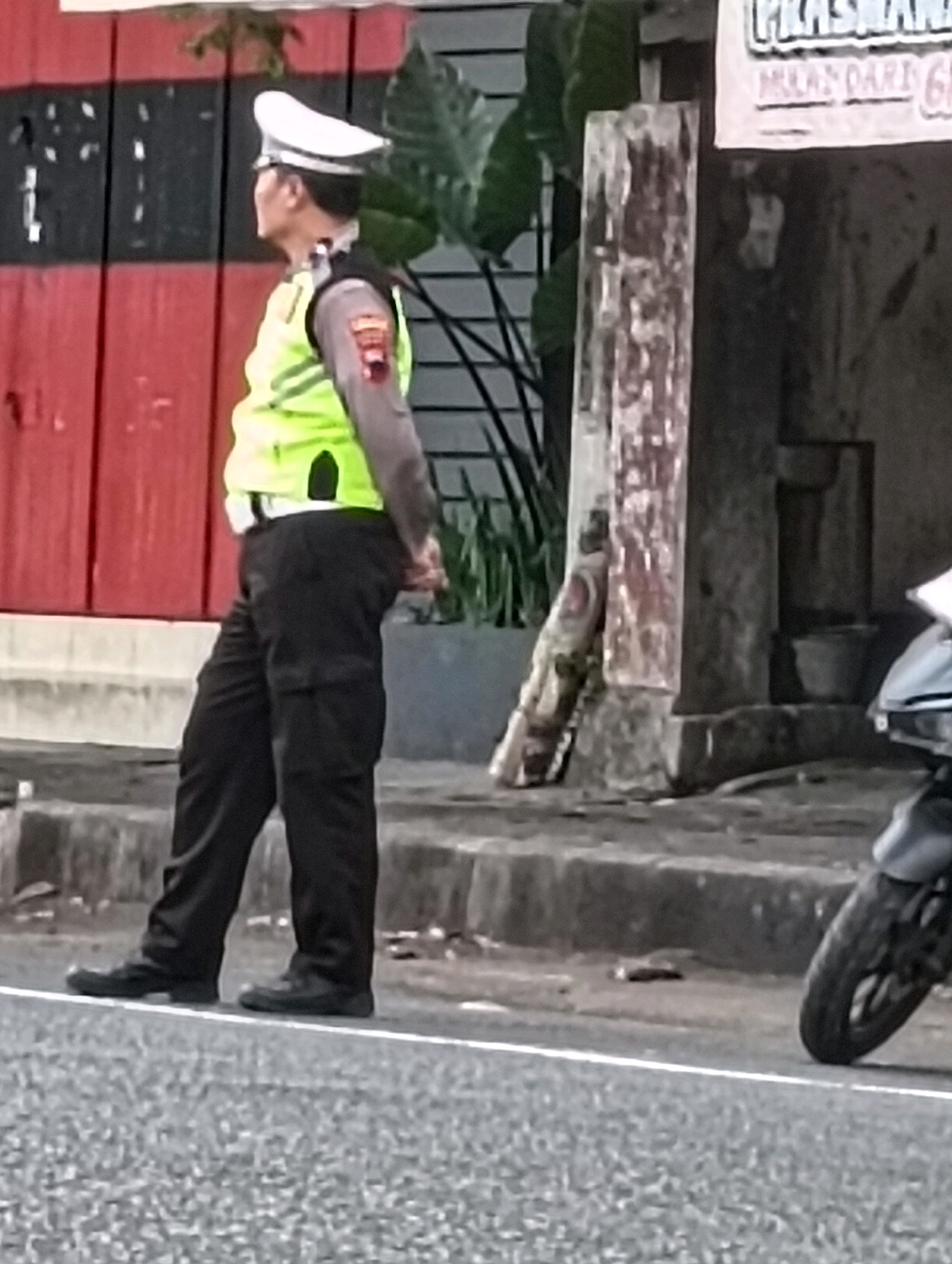The image depicts a police officer in uniform, standing on a sidewalk near a street in Purworejo, Indonesia. The officer is wearing a white cap, a bright yellow reflective vest over a grey uniform shirt with a patch on the left arm, and black pants. The officer's hands are clasped behind their back, and they appear to be looking to the left, away from the camera.
The environment suggests an outdoor urban setting. Behind the officer, there is a building with red and black sections. To the right of the officer, a wooden post with some signage is visible. Further to the right, a motorcycle is partially visible, with its front wheel and handlebars turned to the right. The ground in front of the officer is a paved surface, likely a street, with a white line marking the edge. The sidewalk area where the officer is standing has some concrete elements and loose debris. Lush green foliage can be seen in the background, suggesting the presence of trees or plants. The overall lighting suggests it is daytime.