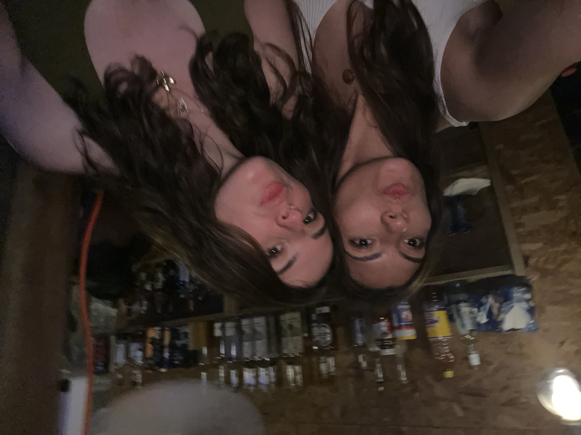 Two women are positioned closely together, with their faces dominating the frame. Behind them is a bar with numerous bottles of alcohol and other items arranged on shelves. The bar appears to be located in a dimly lit area with wooden walls and floors. The location is Puerto Escondido, Mexico.
