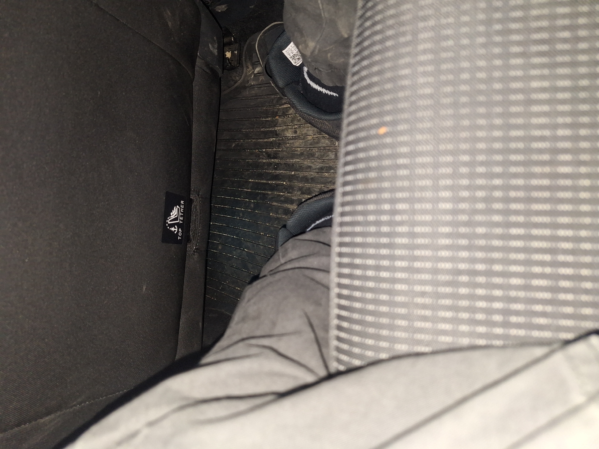 The image shows a car interior. The perspective is from within the car, looking down. A car seat with a grey and white patterned upholstery is visible on the right. To the left is a dark car seat with a "TOP TETHER" tag on it. On the floor, in between the seats, is a black rubber floor mat. A person wearing grey trousers and black shoes with white accents is also visible. No information to indicate if this image was taken in the city of Boliartsi, Bulgaria, exists.