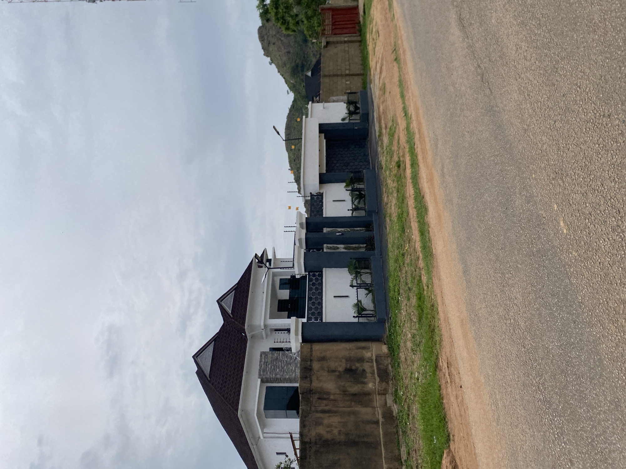 A rotating shot captures a modern, multi-story house situated next to a paved road in Jos, Nigeria. The house features a striking design with a combination of white and dark grey facades, punctuated by large windows and balconies. The roof is a rich, dark brown, with two prominent gables. Part of the house has a stone-clad section, adding texture to the exterior.
In front of the house, a dark grey fence encloses the property, topped with security wiring, suggesting a residential area with security concerns. Some small palm trees are visible within the fenced area, adding a touch of greenery. Beyond the house, a steep, rocky hill dominates the background, covered in sparse vegetation. The sky is overcast, with soft, grey clouds obscuring the sun, indicating a cloudy day.
To the right of the house, the paved road stretches into the distance, with a narrow strip of grass and dirt bordering its edge. The road surface appears to be asphalt, showing signs of wear. There is no visible text in the image. The overall impression is one of a well-maintained, contemporary home in a semi-urban or suburban setting, potentially with a slightly rugged natural landscape nearby. The scene is static, with no discernible activity taking place.