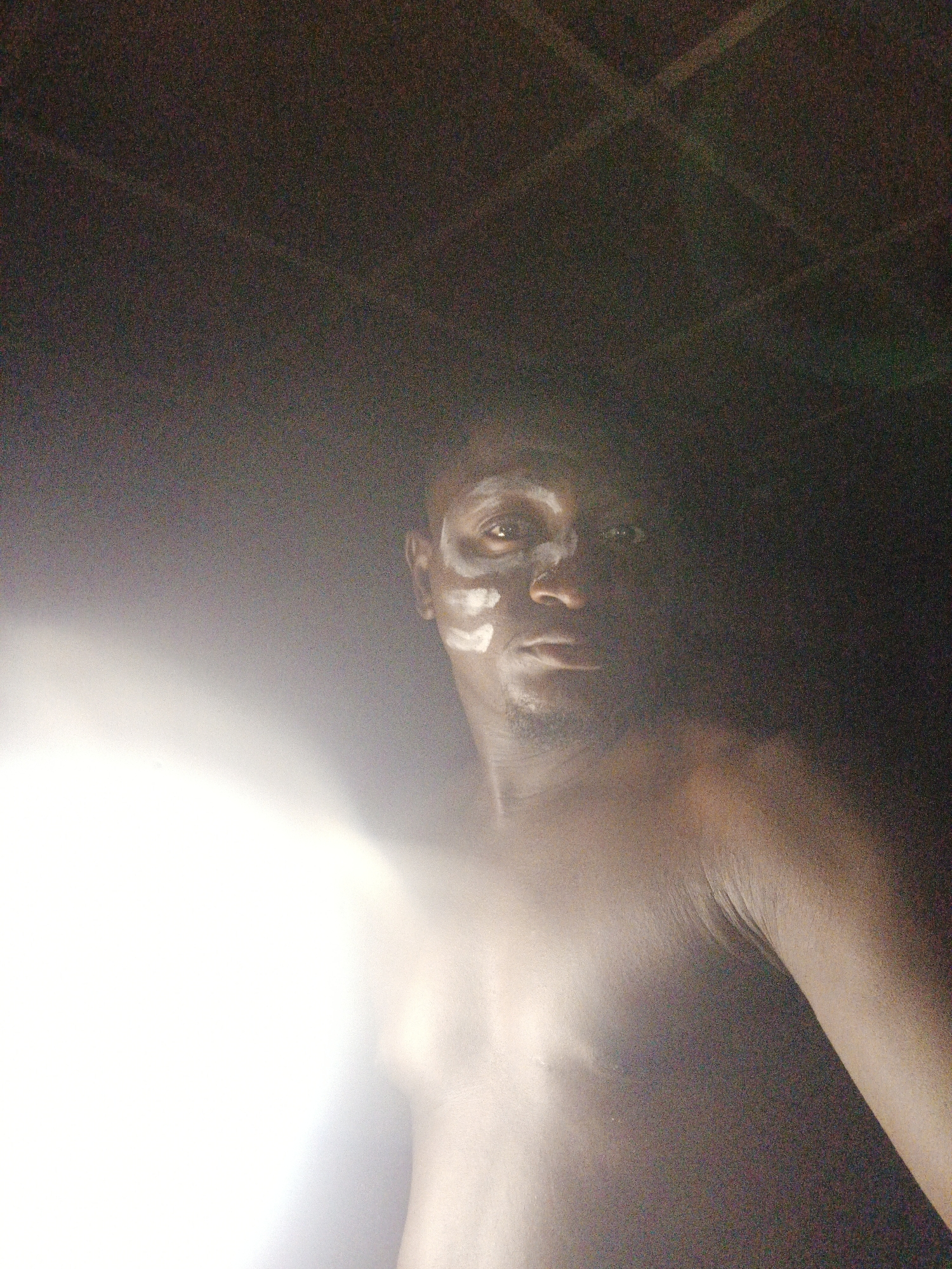 A close-up portrait features a dark-skinned individual, likely male, with white ritualistic or ceremonial markings applied to their face. The markings are visible on the forehead, under the eyes, on the cheekbones, the bridge of the nose, and the chin. The person is shirtless, revealing their chest and right shoulder, and looks directly at the camera with a neutral expression.
The scene is extremely dark, suggesting it was captured at night or in a very dimly lit indoor space. A powerful light source on the left side of the frame creates a significant lens flare and overexposure, strongly illuminating the left side of the person's face and body while casting the right side and much of the background into deep shadow. The background appears dark and grainy with indistinct textures. The high-contrast lighting creates a dramatic and somewhat mysterious atmosphere. The facial markings, combined with the low-light setting, suggest a cultural performance, ceremony, or traditional portraiture, possibly reflecting local customs in Akwanga, Nigeria. No text is visible in the image.