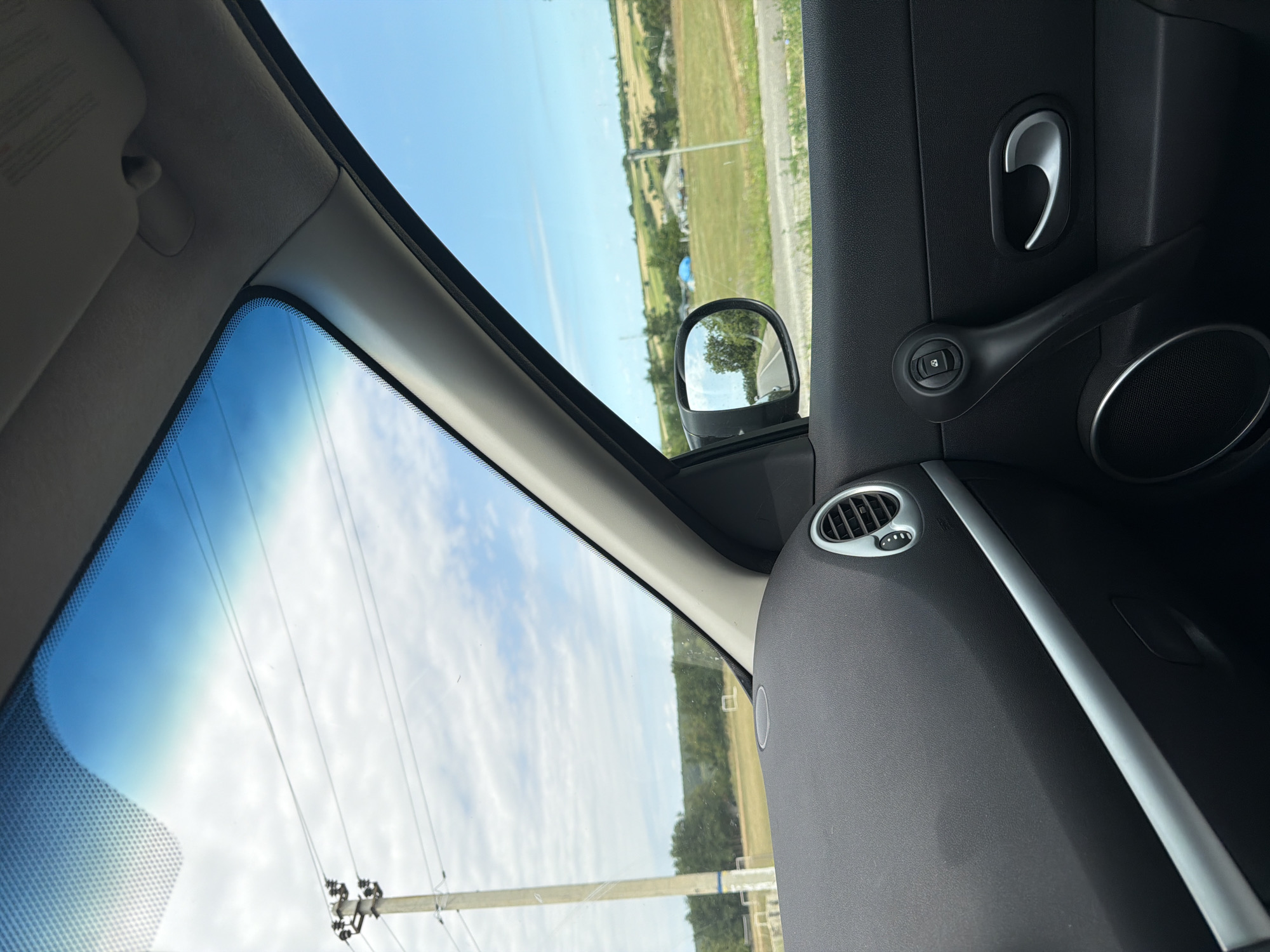 The image, taken from inside a vehicle, provides a view through the windshield and side window. The dashboard and door panel of the car are visible in the foreground, featuring black textured plastic, a silver accent strip, a circular air vent, and a door handle. Through the windshield, a bright blue sky with scattered white clouds is seen, along with a utility pole and power lines. The side window shows a rural landscape with green fields, trees, buildings, and a blue tarp. A side-view mirror reflects the road ahead and trees. The scene is likely situated in or near Rublenița, Moldova, based on the description.