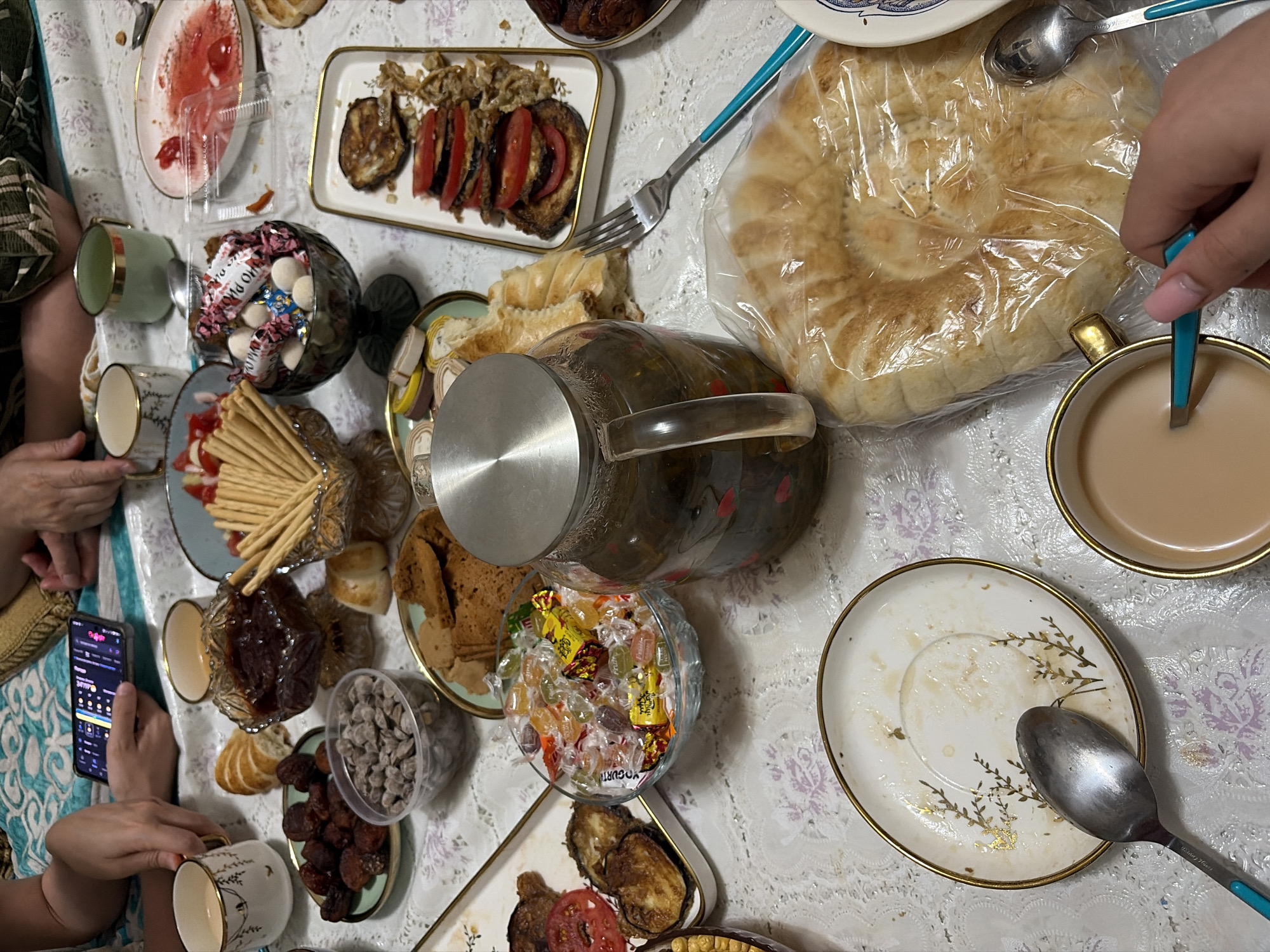 This overhead shot captures a spread of food and drinks on a table, likely a meal or gathering. The setting appears to be indoors, with a patterned tablecloth and glimpses of patterned fabric from clothing. A large, round flatbread, wrapped in plastic, is a prominent object, along with a glass pitcher filled with a dark liquid, possibly tea or juice, and pieces of fruit. Several small cups, some with gold trim, are scattered around, and one contains a light brown beverage, likely coffee or tea. There are also plates holding various snacks, including what looks like fried eggplant with tomatoes, dried fruit, crackers, and a bowl of candies.
The presence of multiple cups and dishes, along with hands reaching for food and drinks, suggests a social event or meal in progress. The lighting indicates it's daytime. One hand is holding a smartphone, displaying a screen with a grid of colorful icons, possibly indicating a casual use of technology during the gathering. The overall mood seems relaxed and communal. There is no visible text that can be read clearly, except for a possible brand name on a spoon which is too blurry to identify. The location context of Astana, Kazakhstan, suggests a meal that might include traditional Kazakh cuisine.