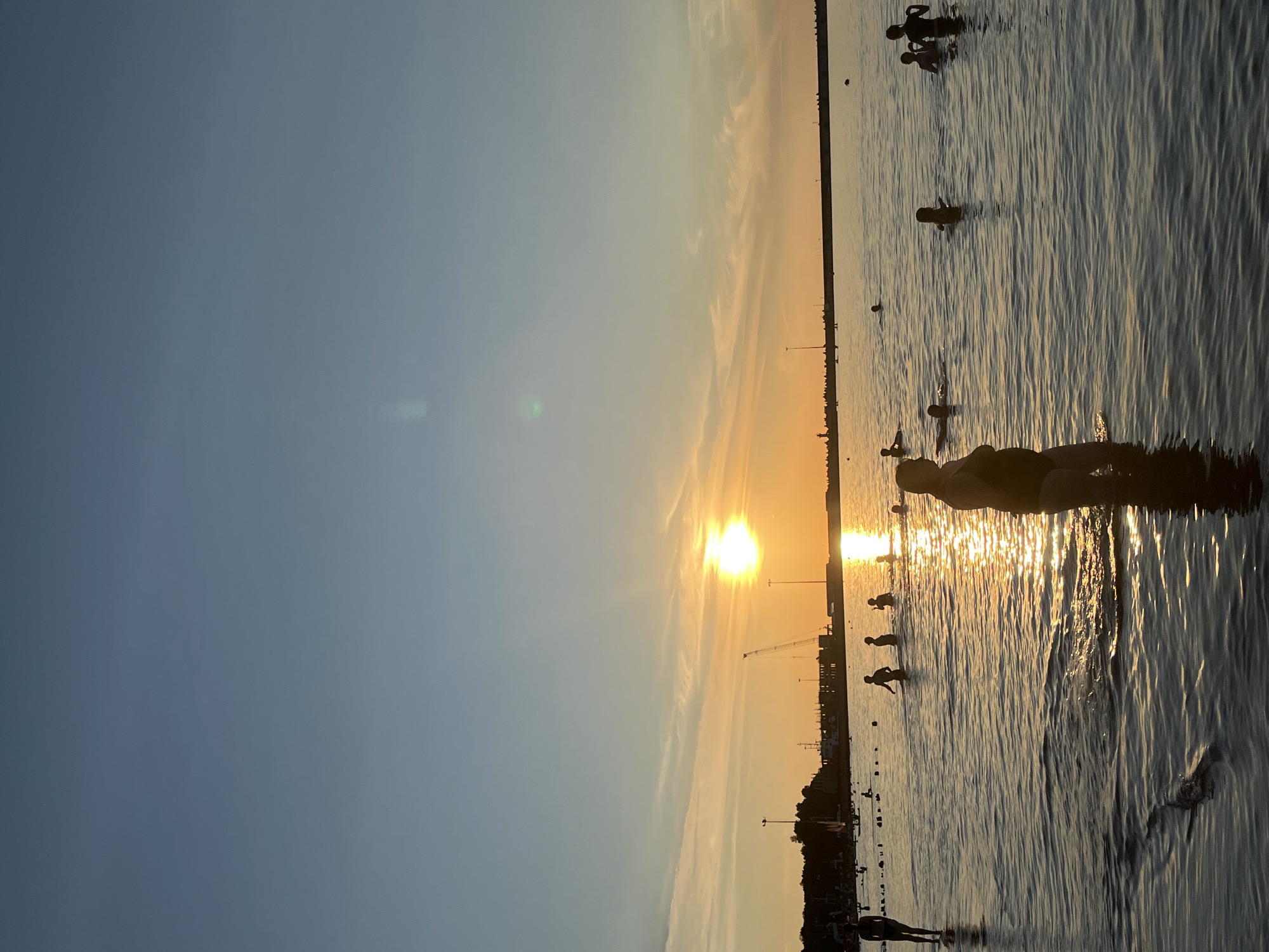 In Pionersky, Russia, on a body of water, people are swimming and wading as the sun sets. The sky is a gradient of blue and orange, with the sun a bright orb near the horizon, casting a golden reflection on the water's surface. Silhouettes of people are visible in the water, some partially submerged, others walking. In the distance, a shoreline is lined with trees and structures, including a tall crane. The overall atmosphere is peaceful and serene, capturing a moment of recreation during a beautiful sunset.