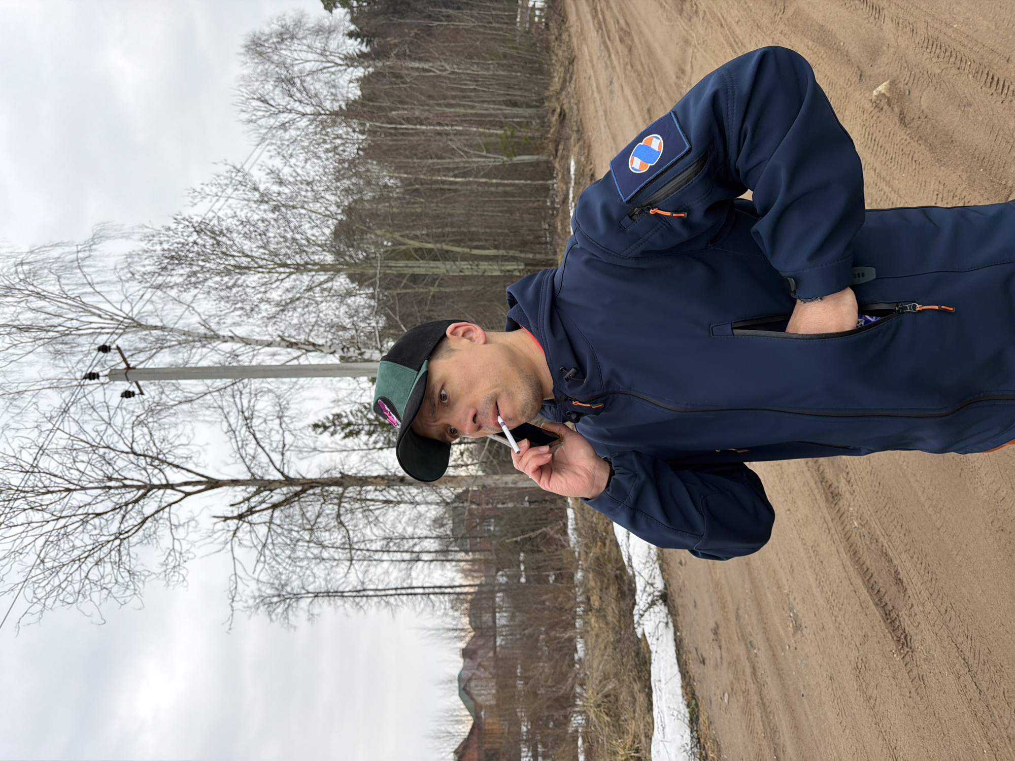 A man in a navy blue jacket with an insignia on the left sleeve and a green and black baseball cap is standing on a dirt road in Chaikovsky, Russia. He is holding a cell phone in his left hand and has a cigarette in his mouth. He is facing left of the frame. The road has tire tracks, and there is some snow along the side of the road. In the background, there are bare trees, a few buildings, and an overcast sky. A power pole is visible to the left of the man. The man has his right hand in the right chest pocket of his jacket.