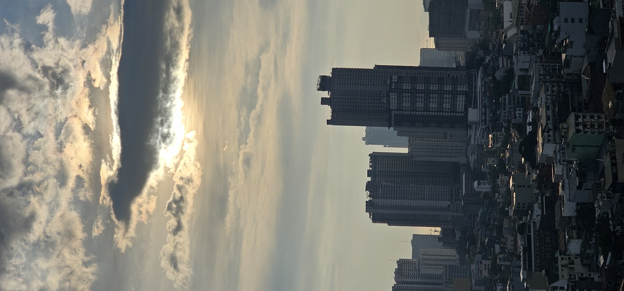 The image captures a cityscape of Makati, Philippines, under a cloudy sky where the sun's rays break through. Dominating the middle ground are several high-rise buildings, some appearing to be under construction with visible scaffolding and unfinished elements. In the foreground, a dense cluster of lower-rise buildings, residential or mixed-use, stretches across the frame, casting long shadows. The overall atmosphere is muted, with the light filtering through the clouds creating a soft, diffuse illumination. No people are visible in the frame. The environment is urban, characterized by a dense built-up area with a mix of modern and older structures.