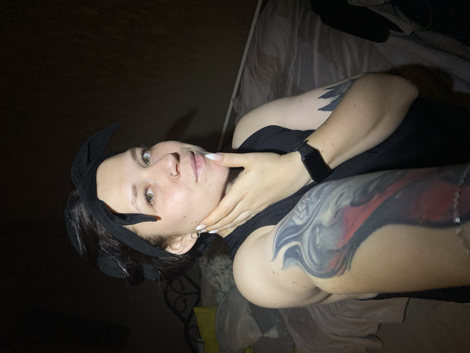A woman with a black bow headband, a nose ring, and ear piercings poses for a selfie indoors in low light. She wears a black top and a smartwatch on her left wrist. Her left arm, visible in the foreground, features a large tattoo. The background includes a bed with gray and white bedding, and a dark wall. The location in the city of Chaikovsky, Russia is unknown, based on the provided image.