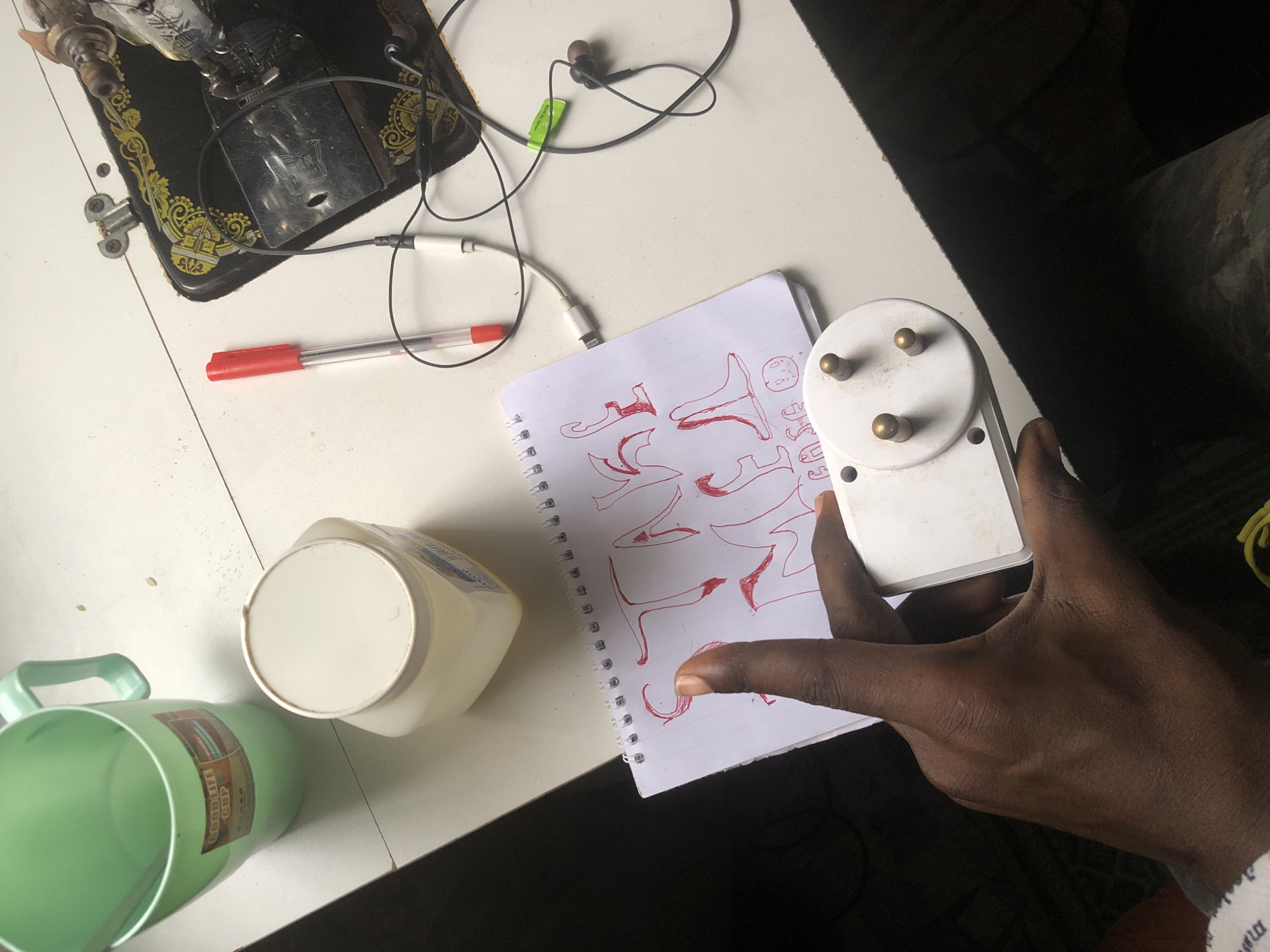 In Ado, Nigeria, a hand of a dark-skinned person is shown holding a white electrical plug adapter with three brass prongs. The adapter is positioned over a spiral-bound notebook with red drawings of letters and a smiley face. The notebook rests on a white surface, possibly a table, which also has a red pen, a pair of black earphones with a green tag, a green plastic cup, and a white plastic container. A sewing machine is partially visible in the background, suggesting a domestic or workshop environment.