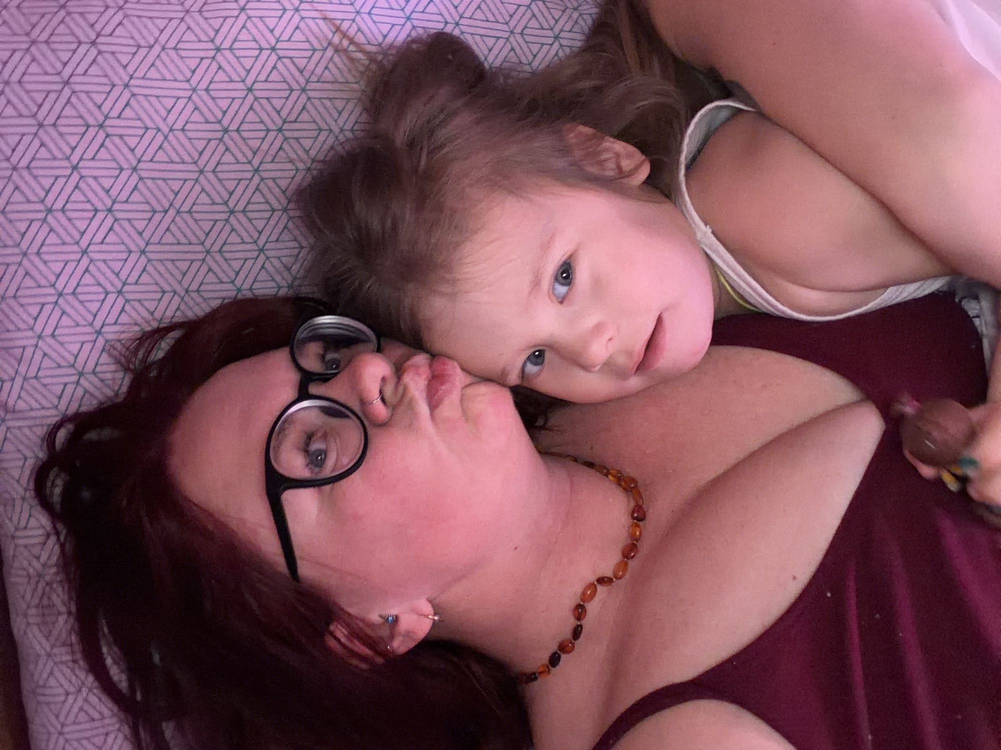 An overhead view captures an adult female and a young child lying together in an indoor setting in Chaikovsky, Russia, consistent with a bedroom. The adult, with reddish-brown hair, wears black-rimmed glasses, a nose ring in her left nostril, earrings in her left ear, and an amber-beaded necklace. She is dressed in a dark red camisole and has her lips pursed in a kissing gesture, directed towards the child. The child, with light brown hair in a bun, has blue eyes and wears a white tank top. The child is looking directly towards the camera with a neutral expression. Both individuals are positioned closely, suggesting an embrace. They are lying on a light pink/lavender surface patterned with repeating geometric lines, indicative of bedding. The lighting in the scene has a slight pinkish hue.