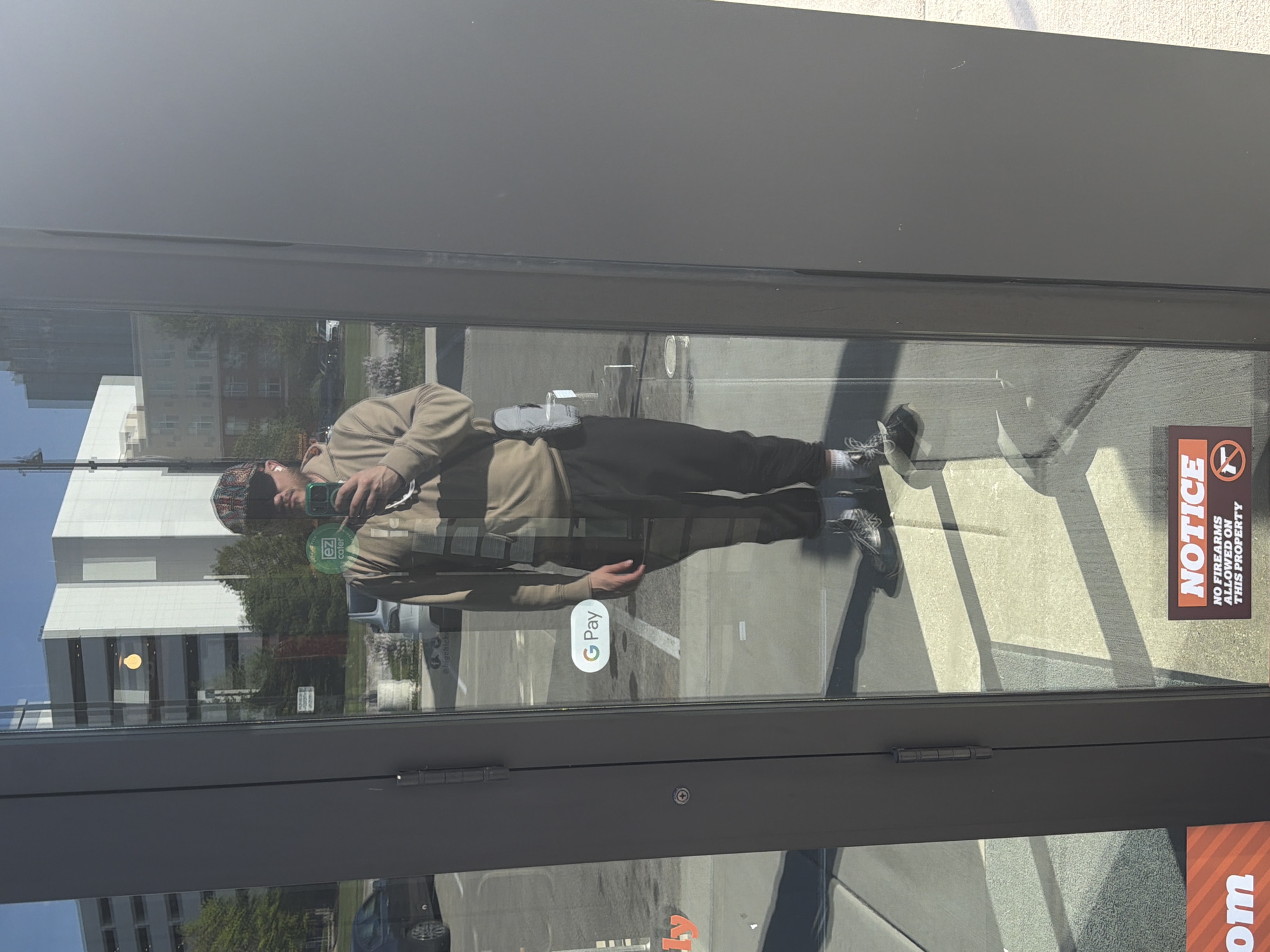 A man is reflected in the glass entrance of a business in Rosemont, Illinois. He is wearing a brown hoodie and dark sweatpants with white socks and gray sneakers. He is wearing a multicolored beanie. He is holding a smartphone with a green circle visible on the back in front of his face, appearing to take a photo of himself in the reflection. The door also has a “G Pay” sticker and a “NOTICE: NO FIREARMS ALLOWED ON THIS PROPERTY” sticker. The reflection in the glass door shows a building, trees, and a car.