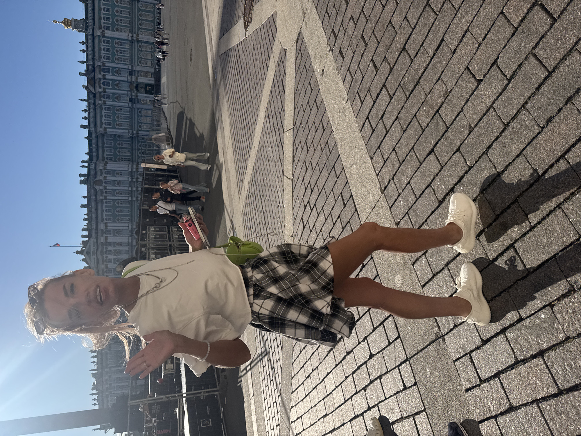 A woman with blonde hair pulled back in a ponytail is walking on a cobblestone street in Palace Square, Saint Petersburg, Russia, facing the camera and waving. She is wearing a white t-shirt, a black and white plaid skirt, and white sneakers. She has a small green purse slung over her shoulder and is holding a pink object in her right hand. In the background, the Winter Palace, a large green and white building, is visible, along with other pedestrians and a large monument. The sun is shining, casting shadows on the ground.
