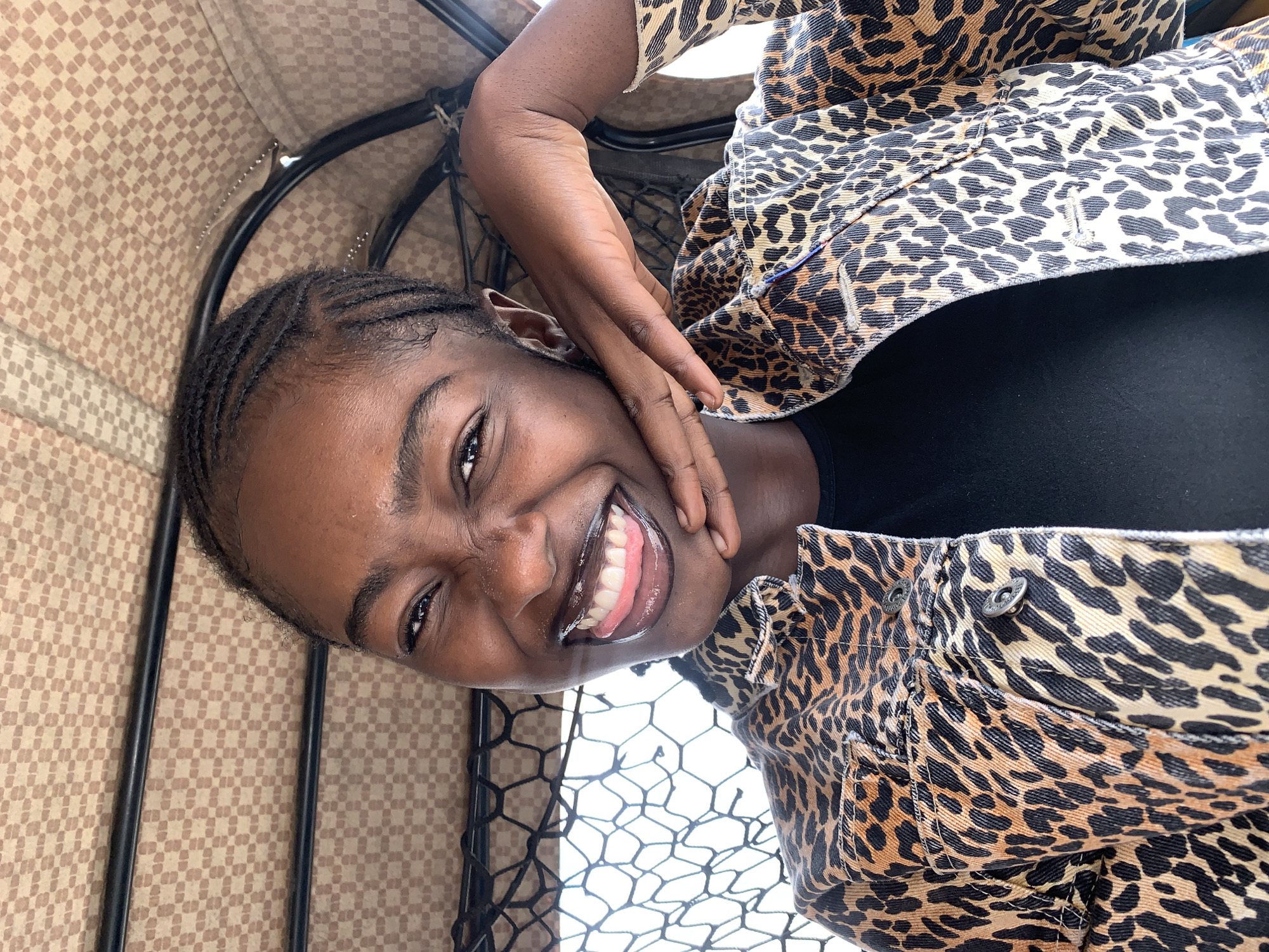 A dark-skinned young woman is captured in a close-up, upward-angled self-portrait within a vehicle, consistent with the interior of a commercial tricycle or auto-rickshaw. She has tightly braided hair styled in cornrows. Her facial expression is a wide, joyful smile, revealing her teeth and with her tongue slightly extended. Her eyes are crinkled at the corners. She is wearing a black top underneath a leopard-print denim jacket, featuring metal buttons on the front placket and pockets. Her left hand is positioned with fingers resting against her chin and jawline. The background features a light brown and beige checkered fabric roof, characteristic of vehicle interiors, supported by black metal bars. A black mesh net is visible to the left, and a bright, unidentifiable exterior environment is glimpsed through an opening. The scene is located in Rafin Par, Nigeria.