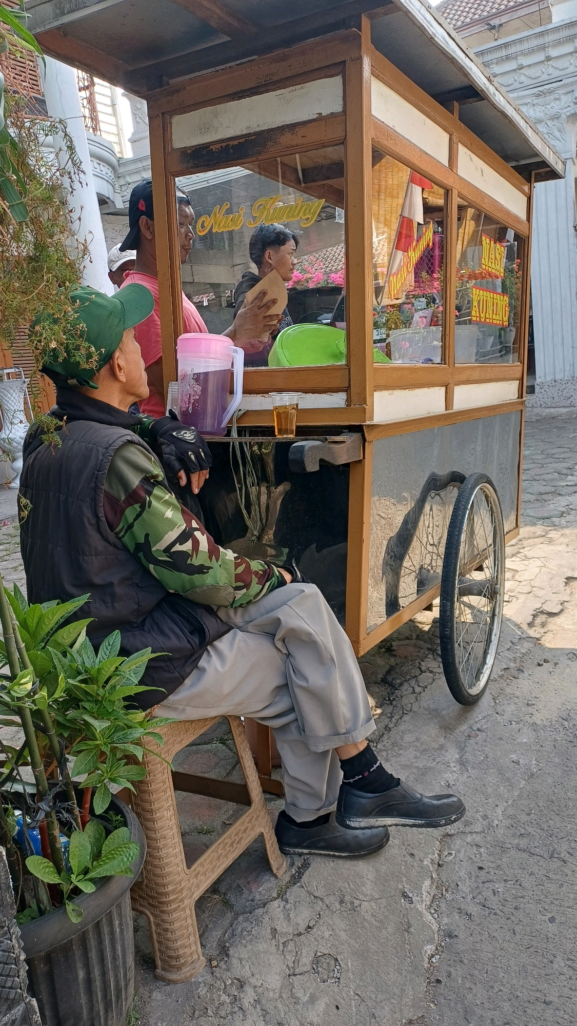 In Gunung Putri, Indonesia, a man wearing a green cap, a black vest over a camouflage long-sleeved shirt, and gray pants sits on a brown plastic stool next to a food cart. The cart, made of wood and glass, displays food items inside, with a sign reading "Nasi Kuning." A pink pitcher and a glass of amber liquid are on the cart. Another man, wearing a pink t-shirt and a black baseball cap, stands behind the cart, looking towards the front. A younger man, also inside the cart, is holding a brown paper item. The scene is set outdoors on a paved surface, with a large potted plant to the left of the man on the stool. The food cart has a spoked wheel on the right side.