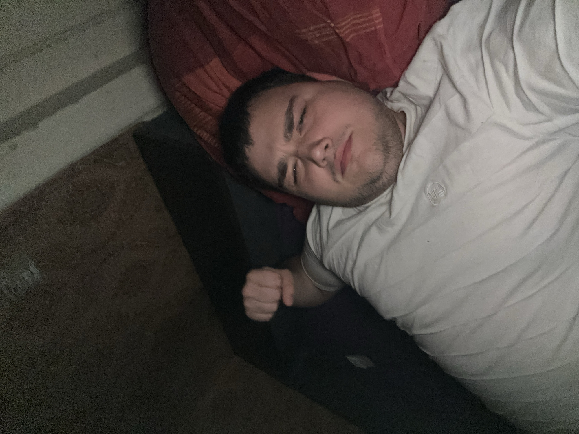 A person is lying on their side on a bed, facing the camera. They are wearing a white t-shirt. Their right arm is bent at the elbow, with their fist clenched near their head. Their eyes are closed and their face is turned slightly away from the camera. The background is dark and out of focus.