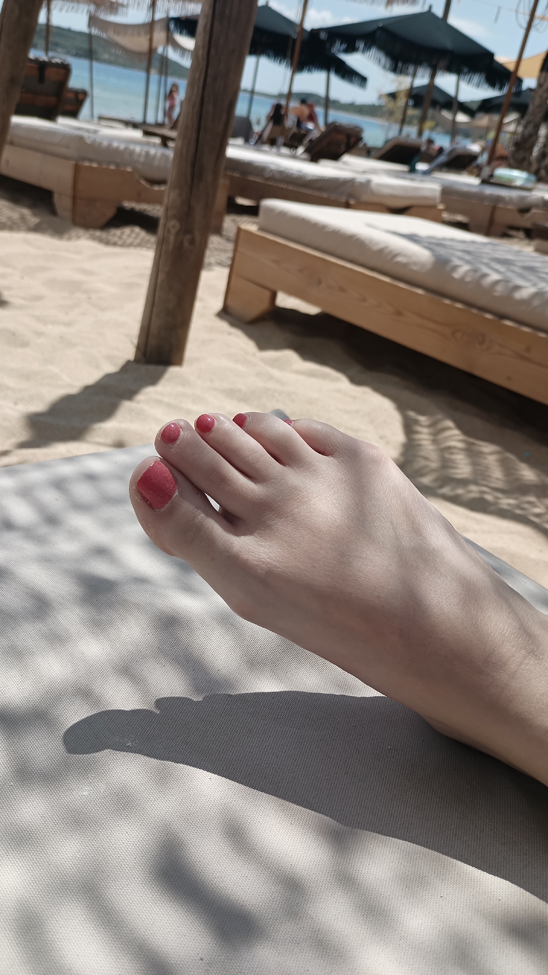 A close-up, low-angle shot focuses on a woman's bare foot, painted with coral-pink nail polish, resting on a white canvas surface. The foot is positioned on the right side of the frame, with toes pointing towards the left. In the background, the sandy beach of Nea Peramos, Greece, is visible, with rows of light-colored wooden sun loungers and beach umbrellas. The sea, a calm blue, is in the distance, with a few people visible on the beach and in the water. Sunlight casts dappled shadows from an overhead structure onto the canvas and the sand.