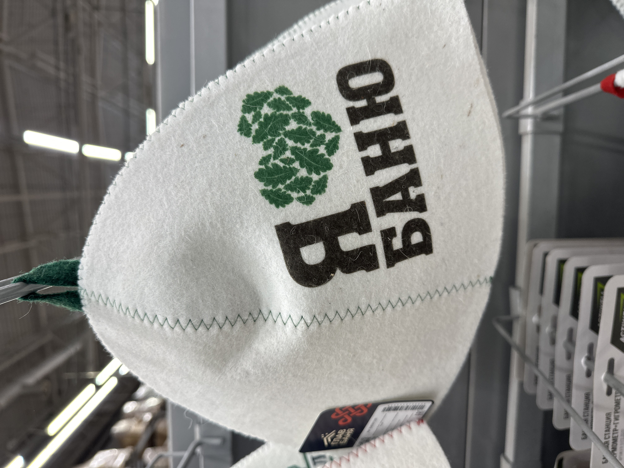 A white, felt sauna hat with green stitching and a green oak leaf heart logo is displayed on a retail shelf. The hat has the Russian text "Я БАНЮ" printed in black, which translates to "I love the bathhouse." The hat is hanging from a metal rod, and in the background, fluorescent lights are visible on the ceiling of a store. The environment appears to be a retail store, possibly a hardware or home goods store, given the packaging of items on the shelves below. The location is identified as Orsk, Russia.