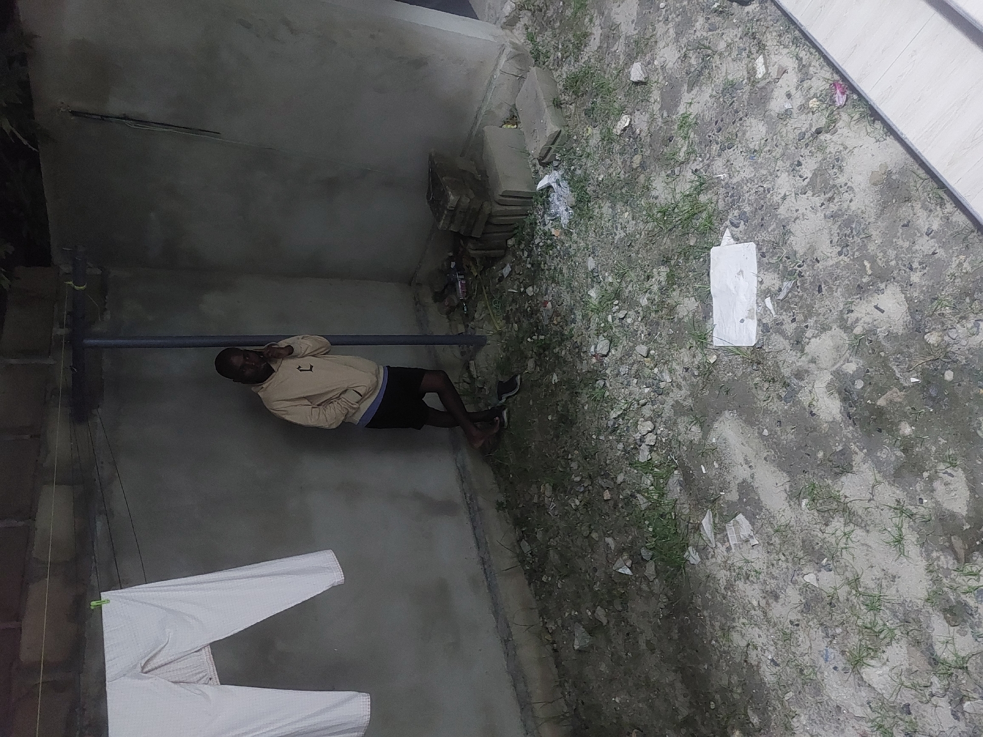 The image depicts an outdoor scene in Dar es-Salaam, Tanzania. A man in a beige jacket and dark shorts lies on a concrete surface. A clothesline with white clothing hangs nearby. The ground is rough with dirt, small stones, and scattered vegetation. A white rectangular object is visible on the ground. The environment suggests a somewhat neglected or less developed area.