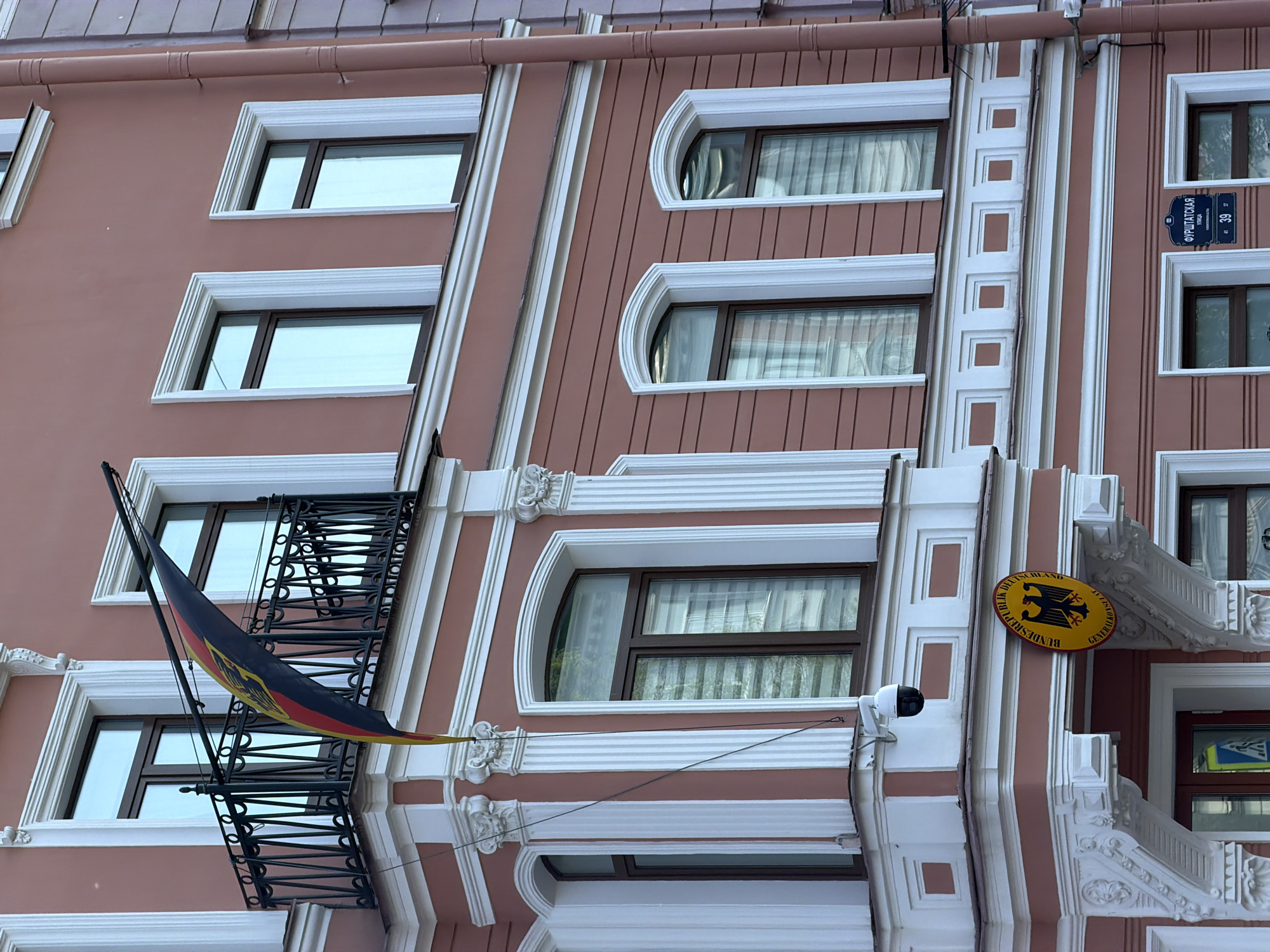 The image shows the exterior of the German Consulate General in Saint Petersburg, Russia. The building has a pink facade with white architectural details. A German flag hangs from a balcony. There is a sign with the German coat of arms on the building. The street sign says "Фурштатская улица", indicating Furstatskaya Street, where the consulate is located. There are several windows with different shapes (rectangular and arched) and white frames. A security camera is visible near the entrance.