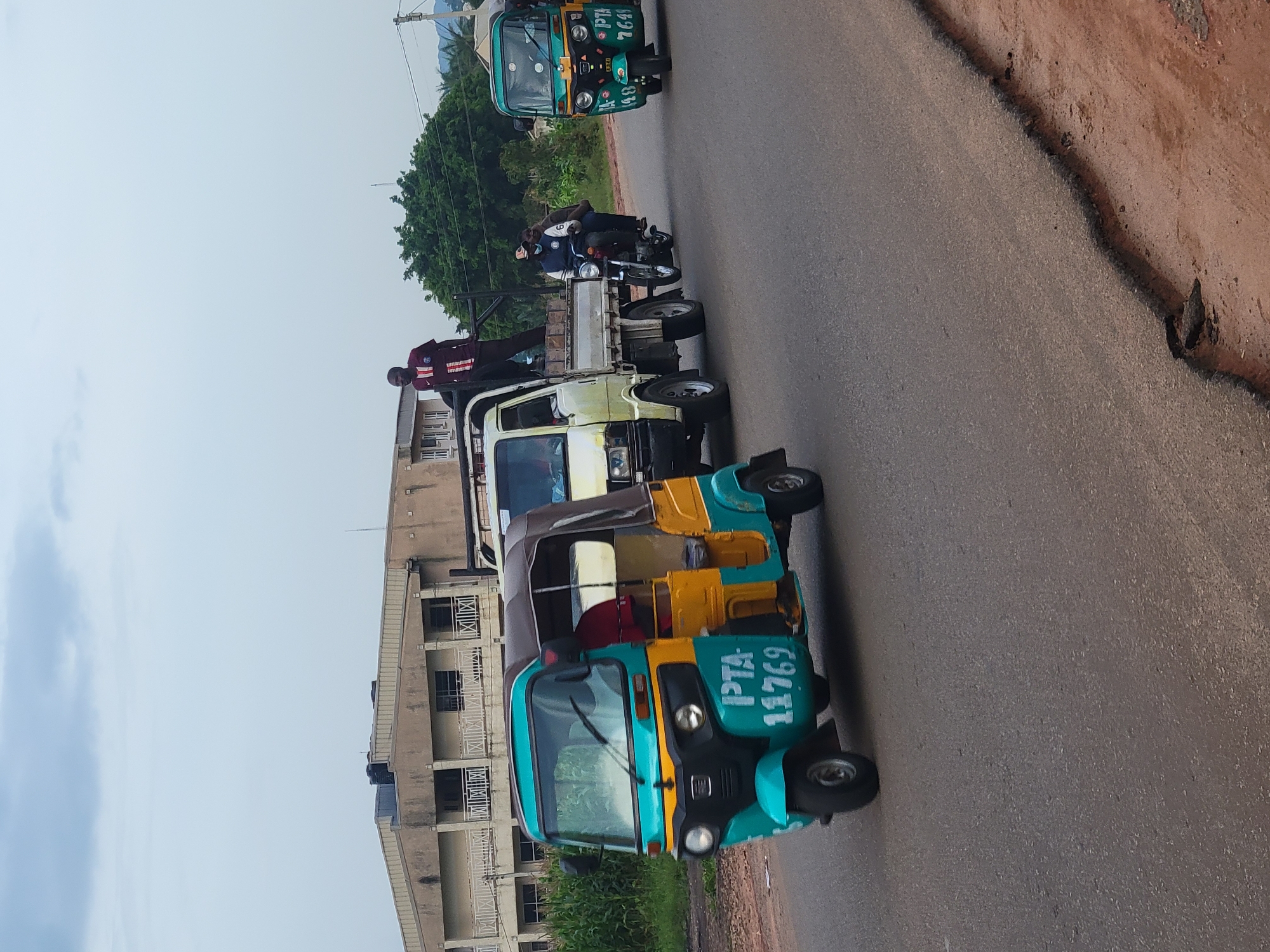 The image depicts a street scene in Jos, Nigeria, likely on a cloudy day. In the foreground, a turquoise and yellow auto-rickshaw, a common mode of transport in Nigeria, is moving on the asphalt road. It bears the registration number "PTA 11769". To its right, a light-colored flatbed truck is also in motion, with a person standing in the bed of the truck wearing a red and white striped shirt. A motorcycle with two occupants is behind the truck. Further to the right, another auto-rickshaw, this one predominantly green, is visible, with the registration "PTA 7641" on its side. In the background, a building with multiple windows and balconies is visible, possibly a residential or commercial structure, with some lush green foliage and trees adjacent to it. The road surface shows signs of wear and tear, with the edge of the asphalt crumbling in places. The overall impression is a busy street with various modes of transportation navigating the road.