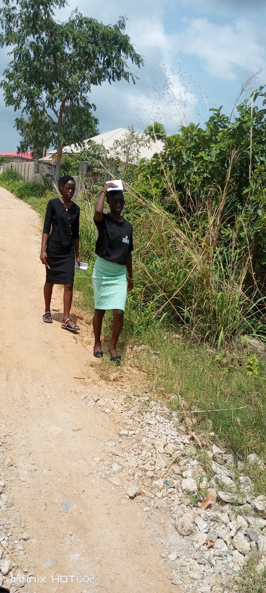 Two young African women walk side-by-side on a dirt road. The woman on the left wears a black dress and sandals. She carries papers in her left hand. The woman on the right wears a black t-shirt, a light green skirt, and flats. She carries papers on her head. They are in Zango-ndaji, Nigeria. There are trees and shrubs on either side of the road. There is a house with a white roof in the background. The sky is partly cloudy.