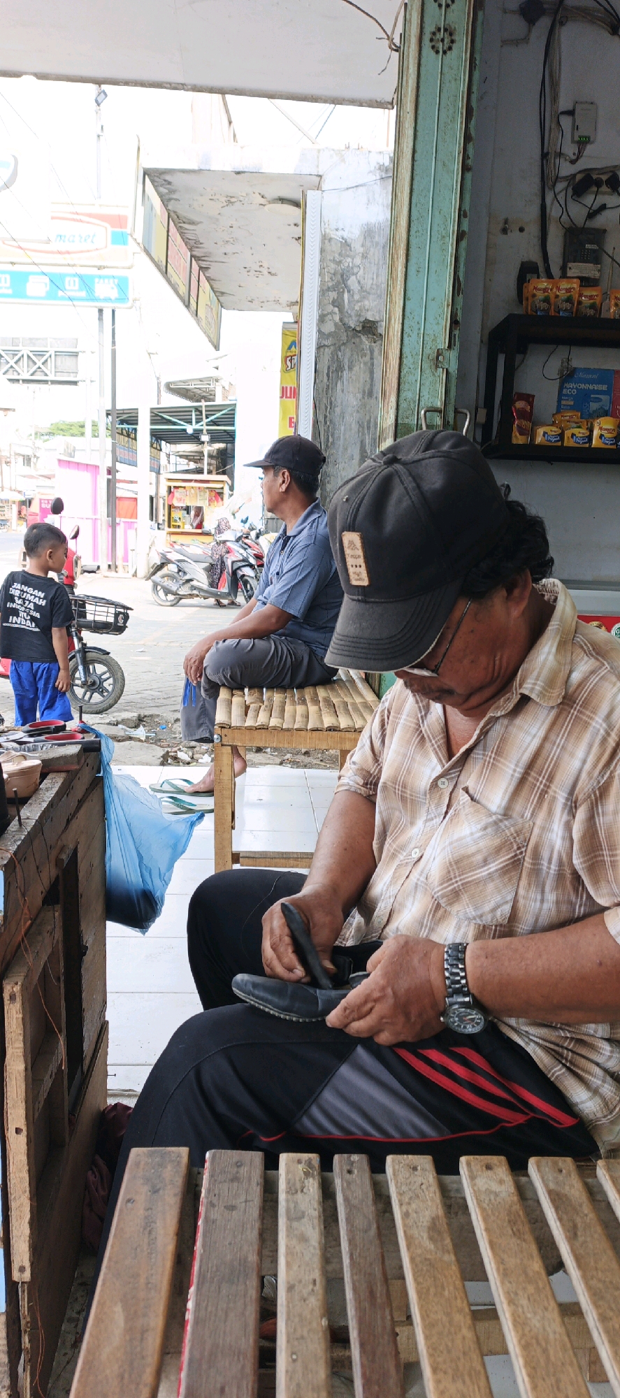 This is a photograph taken in Kasiyan, Indonesia, depicting a street scene outside a shop. In the foreground, a man wearing a plaid shirt, black pants with red stripes, and a black baseball cap is seated on a wooden bench, engrossed in repairing a black shoe with his hands. A wristwatch is visible on his left wrist. To his left, a young boy in a black t-shirt and blue shorts stands next to a motorcycle. In the background, another man in a blue shirt and black cap sits on a similar wooden bench. The shop appears to be open, with shelves stocked with products visible inside. The environment suggests a typical urban setting in Indonesia, with motorcycles and signage indicating commercial activity.
