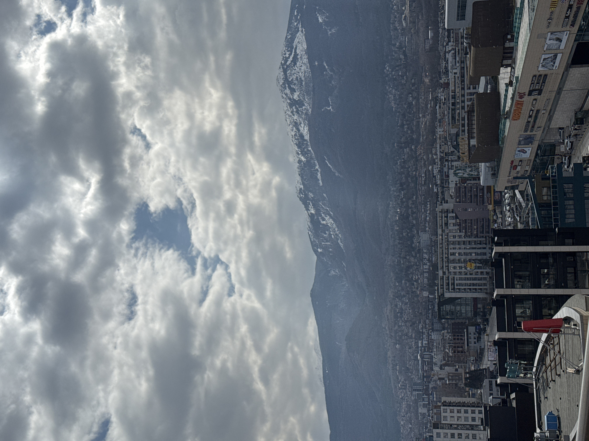 An elevated panoramic view captures a section of Sofia, Bulgaria, with the snow-capped Vitosha Mountain dominating the background under a mostly overcast sky featuring grey clouds and sporadic patches of blue.
The scene depicts a dense urban environment. In the immediate foreground, the rooftop of a dark, multi-story building is visible, featuring a flat, grey surface with various mechanical installations, including blue rectangular units and a prominent red, somewhat cylindrical structure or banner, possibly a windsock or flag, affixed to a metal frame. Adjacent to this, the upper levels and roof of a large commercial building, identified as a shopping center, occupy the right foreground. Its facade displays multiple store logos and names, including "CINEMACITY," "RESERVED," "ZARA," "PULL&BEAR," "BILLABONG," "GUESS," and "DOUGLAS." The roof of this commercial structure supports large, dark, rectangular HVAC units or architectural features.
Below the shopping center, a street runs with several vehicles, primarily cars, and indistinct figures identified as pedestrians. A bus stop structure is also discernible. Beyond these immediate structures, the cityscape extends into the midground with numerous multi-story residential and commercial buildings. The density of construction diminishes as the city merges with the lower, tree-covered slopes of Vitosha Mountain. The mountain's upper regions are distinctly snow-covered, while its forested lower slopes are partially obscured by atmospheric haze. No specific individuals are in focus or performing distinct actions beyond implied pedestrian and vehicle movement.