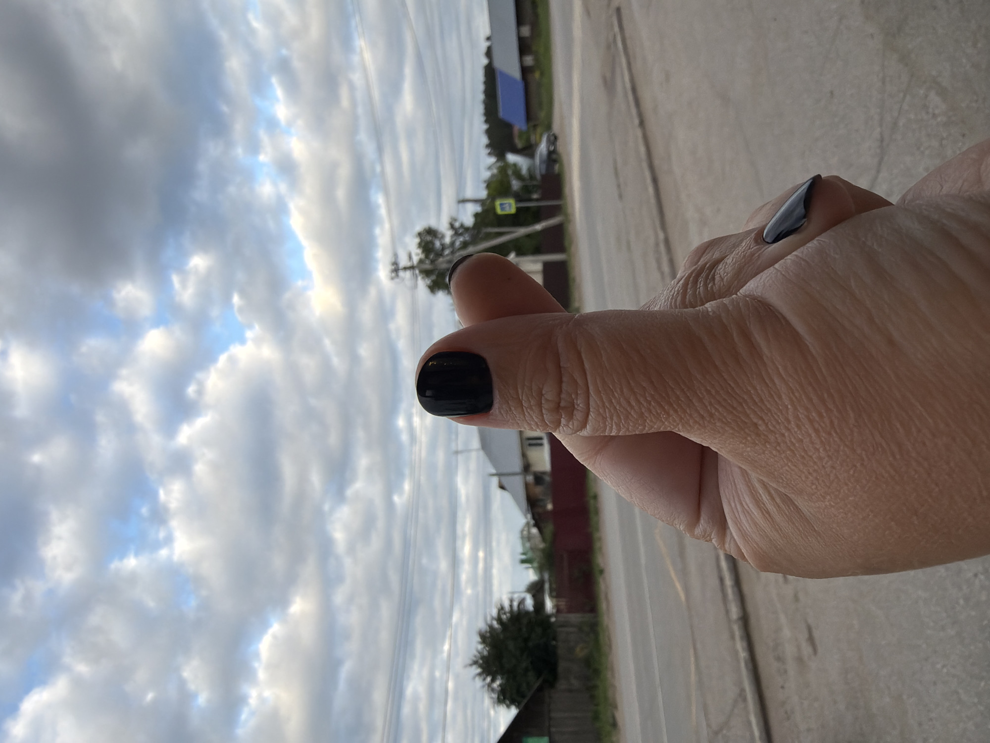 The image shows a close-up of a hand with black nail polish, making a finger heart gesture. In the background, a rural street scene is visible. There are houses with red and blue roofs, a road with a pedestrian crossing sign, and trees. The sky is cloudy. The location is likely Chaikovsky, Russia, given the architectural style of the houses and the presence of power lines, which are common in Russian rural areas.