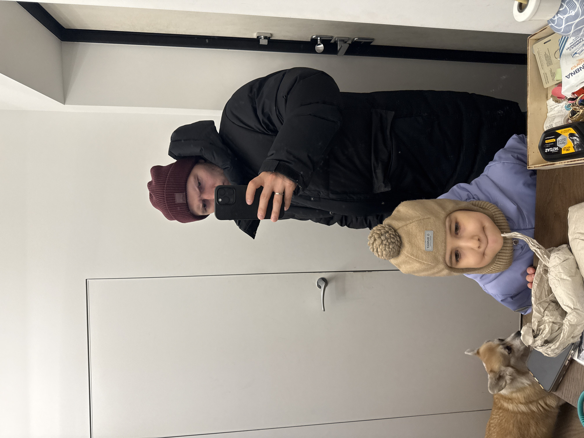 A person in a black puffer jacket and maroon beanie is taking a selfie in a mirror. A child wearing a tan hat and lavender jacket is beside them. A corgi dog looks up at the child. The reflection appears to be in a bathroom with white walls and a drawer unit. A box of small items is on a surface next to the mirror. This scene likely happened inside of a residence, since this photo doesn't show any details that point to the exact location within Krasnoyarsk, Russia.