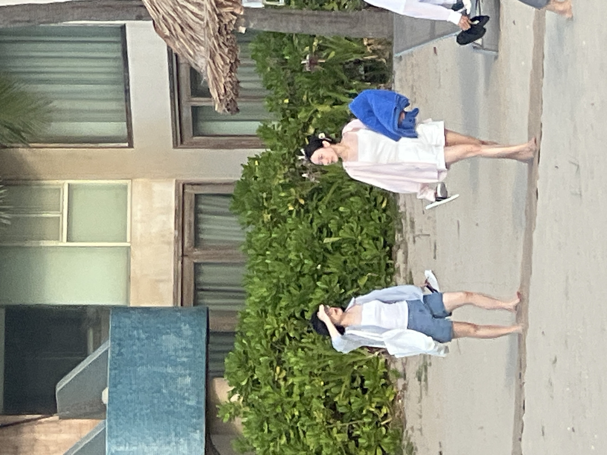 Two women are walking on a sandy beach in front of lush green foliage and a resort building in Đà Nẵng, Vietnam. The woman on the left is wearing a light blue long-sleeved shirt over a white tank top and blue denim shorts. She carries a white tote bag and is shielding her eyes from the sun with her hand. The woman on the right is wearing a white slip dress with a pale pink cardigan and carries a folded blue towel. She is holding a pair of white sandals in her other hand. A thatched umbrella and lounge chairs are visible in the background, suggesting a resort setting. The overall atmosphere is casual and relaxed, typical of a beach vacation.