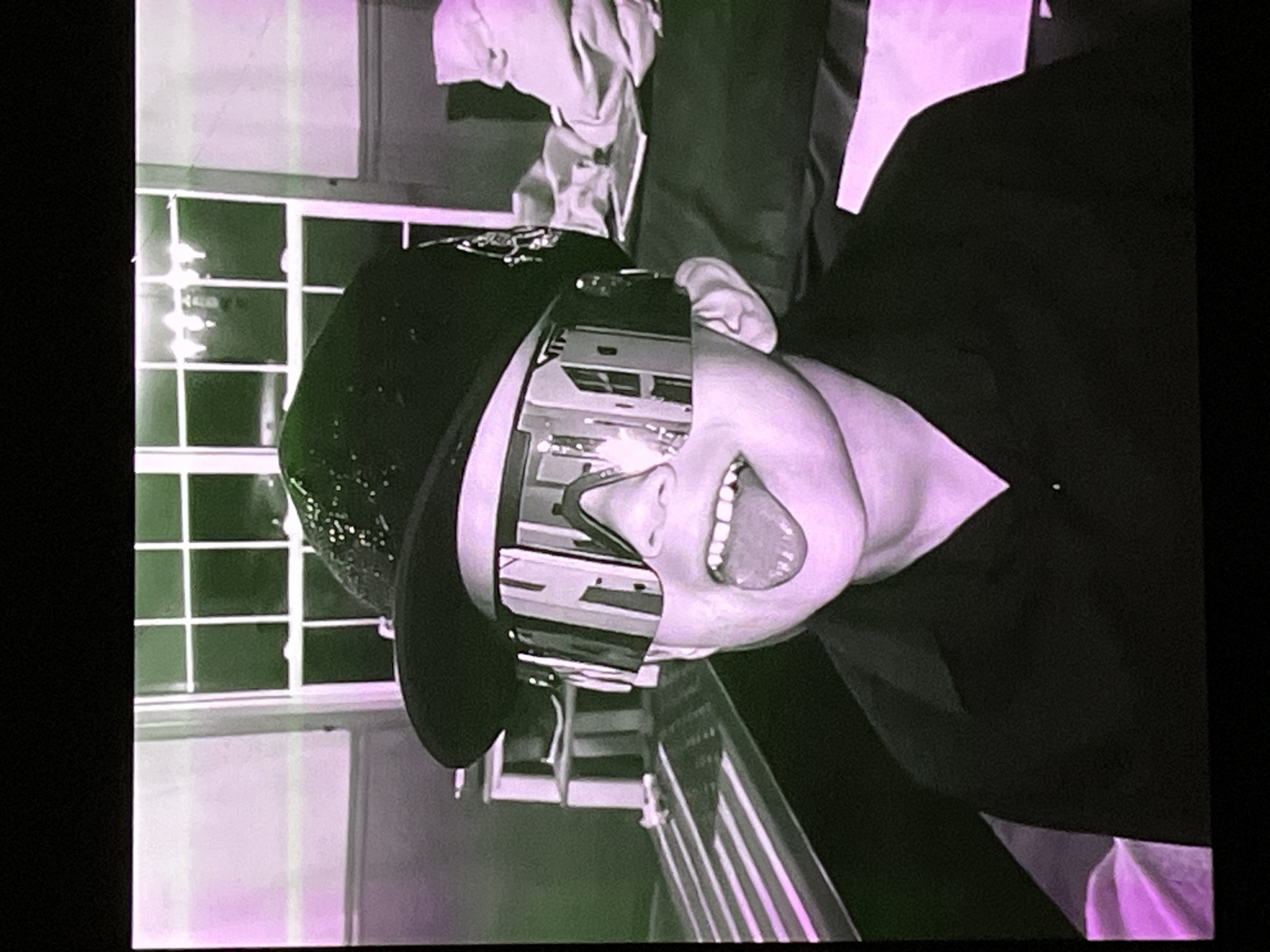 The image shows a young person wearing a black baseball cap with a sparkly pattern and oversized reflective sunglasses. The person is sticking out their tongue and appears to be smiling. They are wearing a black collared shirt. In the background, there is a window with multiple panes, a light fixture hanging from the ceiling, and some shelves with items on them. To the left of the person, there is a striped surface that resembles a table or a race track. To the right, there is a pile of fabric, possibly clothing or bedding. The image has a pink and green color cast. There is no information within the image to suggest its location in Cadek, Indonesia.