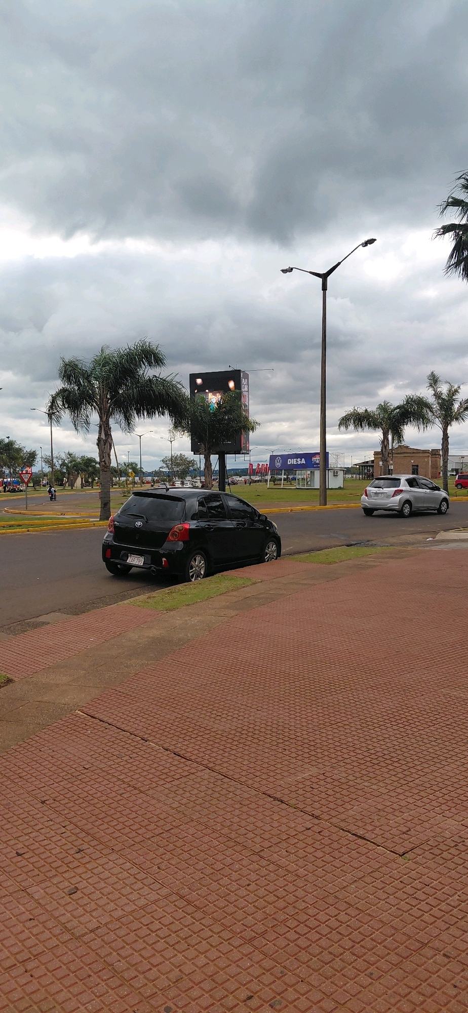 A black Toyota Yaris hatchback is parked on the side of a street in Encarnación, Paraguay, with its front facing away from the camera. The car is on the right side of the road, parked just off the curb. The street has a yellow line marking the edge of the road and a grassy area between the road and a red brick sidewalk. The sidewalk is in the foreground of the image, and it appears to be made of textured concrete tiles. In the background, there is a silver Toyota Yaris driving on the road, and a large digital billboard is visible behind it. There are also palm trees and streetlights visible. The sky is cloudy and overcast.