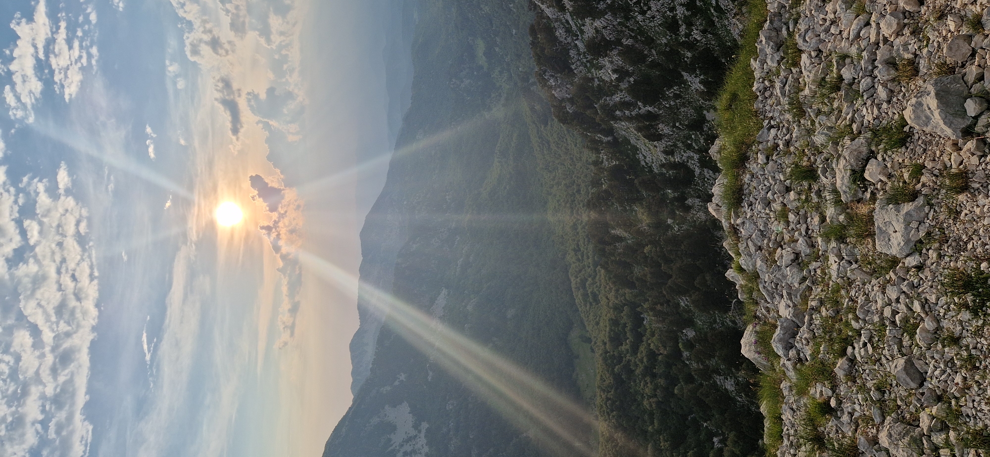 The image captures a breathtaking mountain landscape at sunset, likely in the vicinity of Ubla, Bosnia and Herzegovina. The sun, a bright orb partially obscured by clouds, casts dramatic rays of light across the scene. In the foreground, a rocky terrain is covered with sparse green grass. Beyond this, a lush, verdant valley unfolds, filled with dense trees. Further in the distance, rolling hills and mountains fade into a hazy, atmospheric perspective, bathed in the soft, golden light of the setting sun. The sky is a canvas of soft blues and whites, with scattered clouds that catch the sunlight. The overall impression is one of serene, natural beauty and vastness.