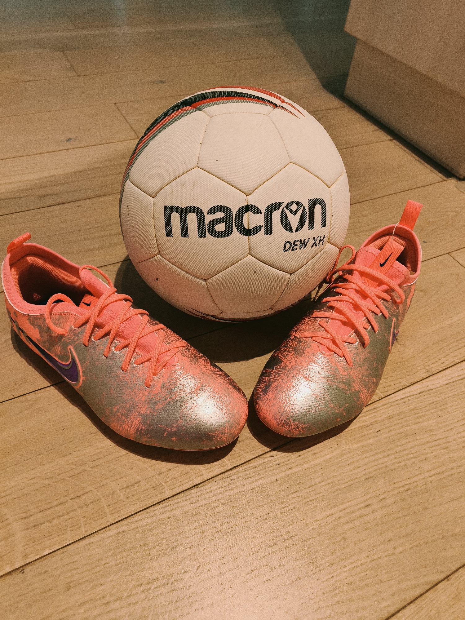 The image shows a white Macron brand soccer ball with red and black stripes on top, and a pair of coral-colored Nike soccer cleats with silver accents and purple swooshes, all placed on a wooden floor. The photograph appears to be taken indoors under warm lighting. The location of the city of Sofia, Bulgaria is not discernible from the image.