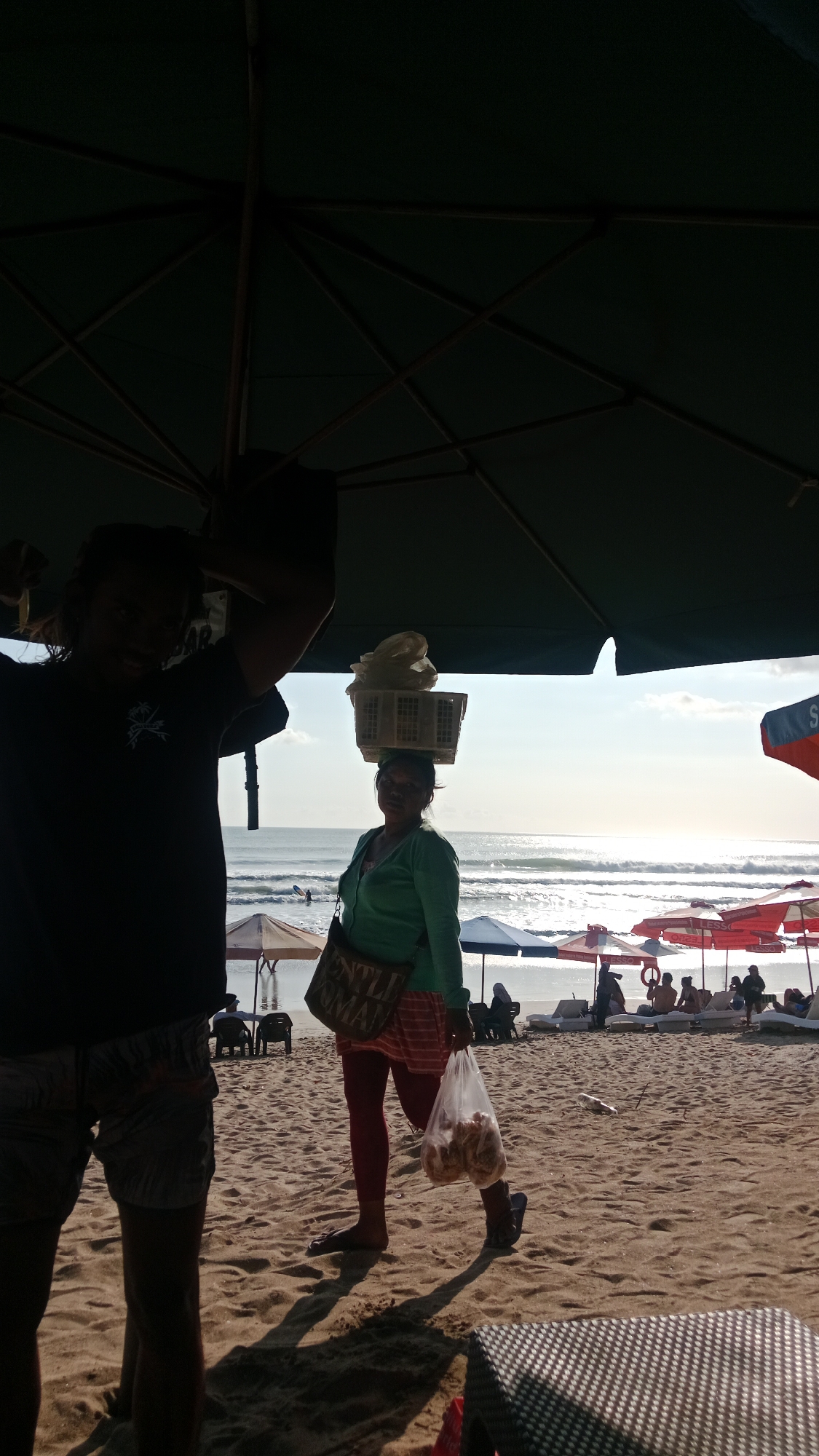 On a sandy beach in Kuta, Indonesia, two figures are partially obscured by a large, dark green umbrella. To the left, a person in silhouette stands with their arms raised, wearing a dark t-shirt and patterned shorts. To the right, a woman walks carrying a basket of goods on her head and a plastic bag of items in her right hand. She is wearing a green long-sleeved shirt, a striped skirt, and dark leggings. The ocean with gentle waves is visible in the background, along with several beach umbrellas and lounging chairs occupied by other beachgoers. The sun is low in the sky, casting long shadows across the sand.