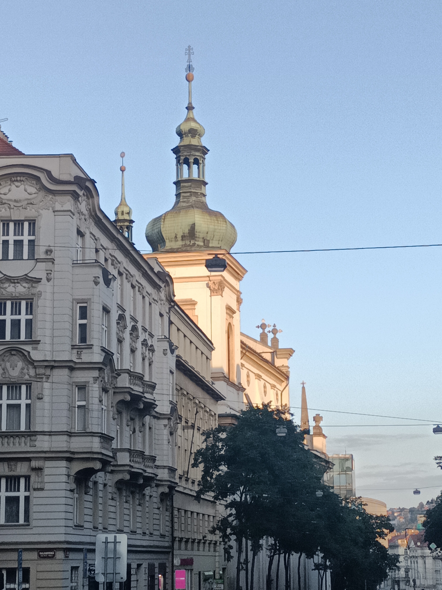 The image shows a street scene in Prague, Czechia, with the St. Nicholas Church's tower visible in the background. The foreground features an ornate grey building with multiple stories and windows, balconies, and decorative architectural elements. A large, dark green tree stands in front of a building to the right, partially obscuring a church spire. Overhead, power lines and what appear to be cable car lines crisscross the sky. In the distance, a cityscape of buildings stretches across rolling hills under a clear, pale blue sky.