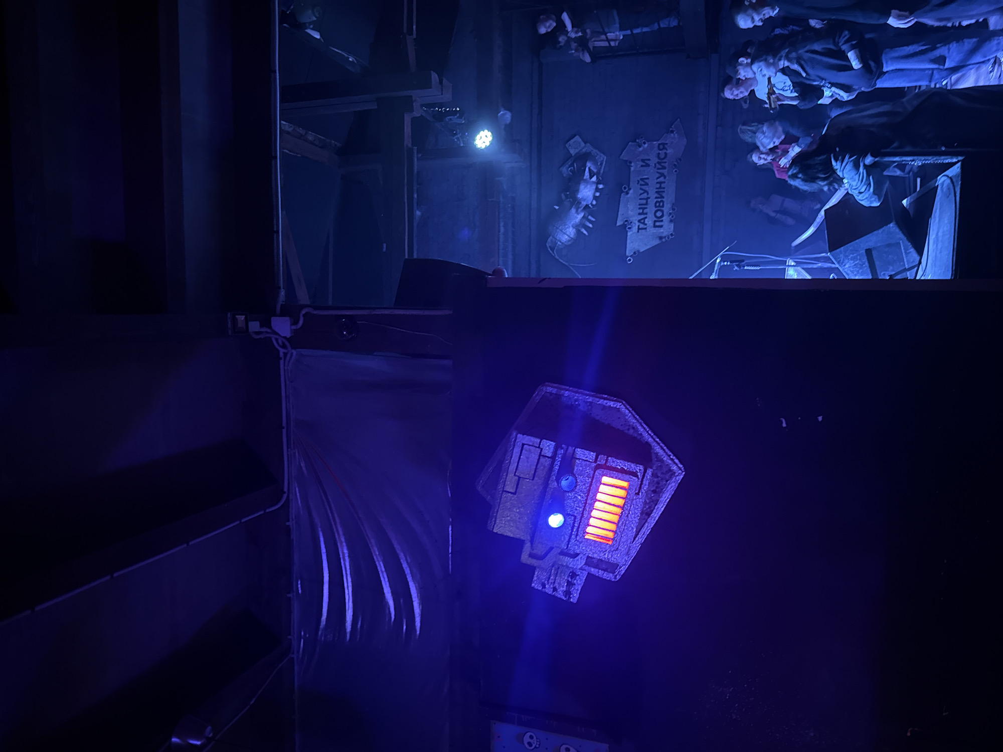 The image shows an interior view with a focus on stage decorations. At the top of the image, there's a glimpse of a crowd of people, likely audience members. Just below them, stage equipment such as speakers and a microphone stand are visible.
On the wall, there's a sign with text in Russian. An illuminated, sci-fi-themed light fixture, displaying a blue and orange light is mounted on the wall. To the left of the light fixture, there's a section of wall with what appears to be a curtain or some draped material. The overall lighting is predominantly blue, creating a theatrical or atmospheric effect.
Without any explicit mention in the provided image, the location in the city of Saint Petersburg, Russia, cannot be confirmed.