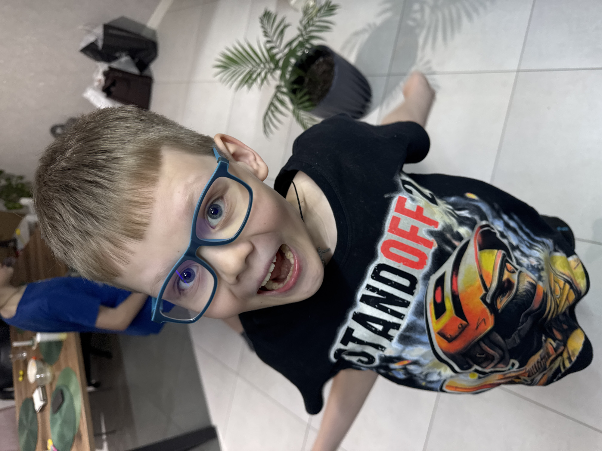 This media file captures a young boy indoors, presented from a high-angle perspective and rotated clockwise. The boy, who has short light brown hair, blue eyes, and blue-framed glasses, is looking directly at the camera with an excited, open-mouthed expression that suggests playfulness or surprise.
He is wearing a black short-sleeved t-shirt featuring a prominent, colorful graphic. The graphic depicts an armored figure or robot in shades of orange, yellow, black, and grey. Above this design, the text "STAND OFF" is clearly visible in white and red lettering.
The setting appears to be a home environment with light-colored square tiled flooring. In the background, to the boy's right, a potted plant with palm-like leaves casts a shadow on the floor. To his left, another individual wearing a blue shirt is partially visible, seated at a wooden table. On the table, several green circular placemats and other household items, possibly cups, are arranged. Further back, some stacked boxes or items are partially seen. The lighting suggests an indoor daytime setting. The overall impression is one of a cheerful and spontaneous moment in a domestic space in Ферма, Russia.
