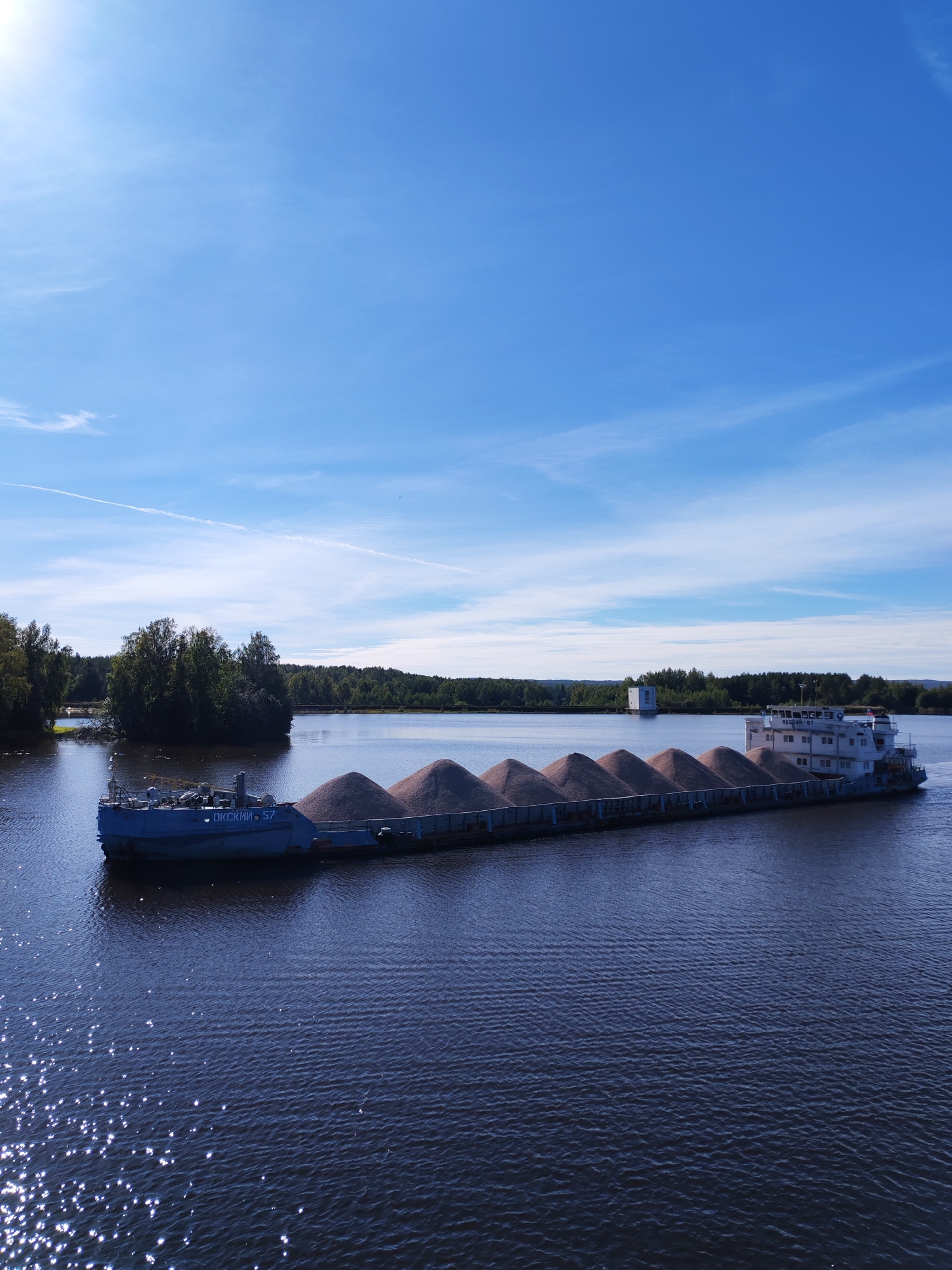 The image depicts a wide, dark blue body of water, likely a river or lake, under a bright blue sky with scattered thin white clouds. A long, flat-bottomed barge is the prominent subject, laden with a cargo of what appears to be sand or gravel piled high in a series of humps. The barge is divided into two sections: a forward, blue-painted tugboat with some visible machinery and a nameplate reading "ОКСКИЙ 57", and a larger, white-painted cargo section towards the stern. The cargo section has multiple decks or cabins. In the background, a lush green shoreline with dense trees lines the water. A small, isolated island covered in trees is visible on the left. Further in the distance, a white, rectangular structure, possibly a building or tower, stands on the far bank. The sun is shining brightly, creating sparkling reflections on the water's surface. The overall scene suggests a clear, sunny day in a natural, possibly rural or semi-rural, environment. The presence of the barge indicates a transport or shipping activity. The location is stated to be in Nikol'skaya Gora, Russia.