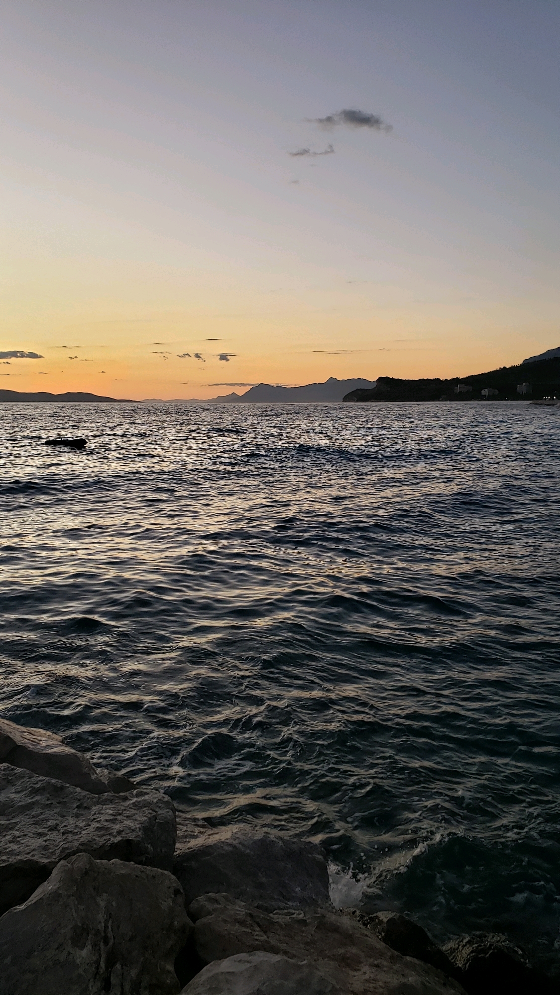 This image depicts a serene sunset over the Adriatic Sea, likely captured from the shore of Općina Tučepi, Croatia. The sky transitions from a soft orange hue near the horizon to a pale blue overhead, with a few wispy clouds present. In the distance, silhouetted mountains form the coastline, with hints of buildings visible on the slopes. The sea is a deep blue, with gentle ripples creating a textured surface that reflects the fading sunlight. Large, grey rocks are visible in the foreground, bordering the water's edge. There are no people or animals visible in the frame, and no specific actions or interactions are taking place. The overall atmosphere is one of tranquility and natural beauty.