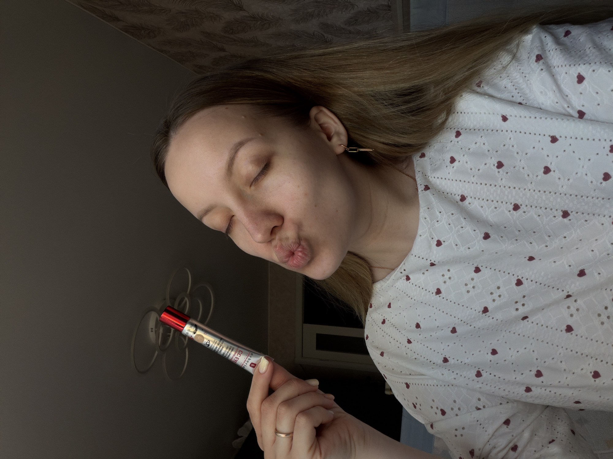 A woman with closed eyes and puckered lips poses with a tube of Erborian CC Eye cream. She has blonde hair and is wearing a white shirt with red heart patterns. She is also wearing a gold ring on her finger and gold, rectangular earrings. The background appears to be a room with a dark wall and a decorative lighting fixture on the wall. There is no information in the image to indicate the location is in Reshetnikovo, Russia.