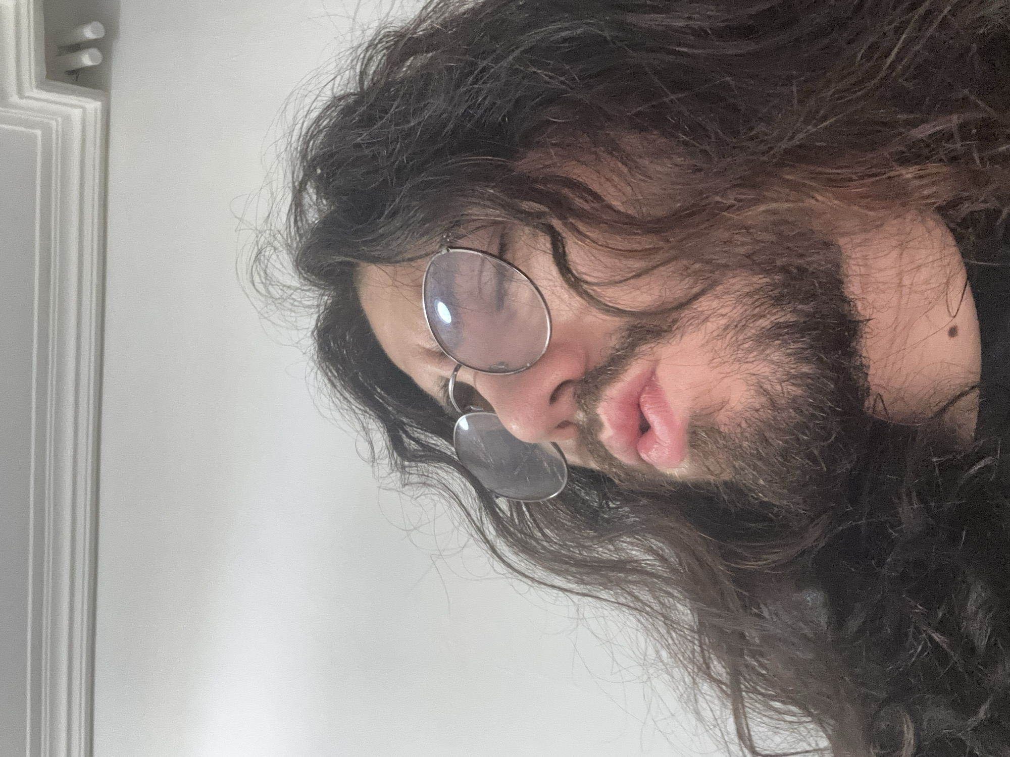 A low-angle, close-up shot focuses on the face of a person with long, dark, messy hair. They are wearing round, wire-rimmed glasses and have a beard. Their lips are pursed, and their eyes are closed, with a reflection visible in the glasses. The background is a plain, light-colored wall with ornate white crown molding at the top. The lighting is bright, creating highlights on the glasses and casting some shadows. The overall impression is a candid, possibly playful, or pensive moment. The scene is set indoors.