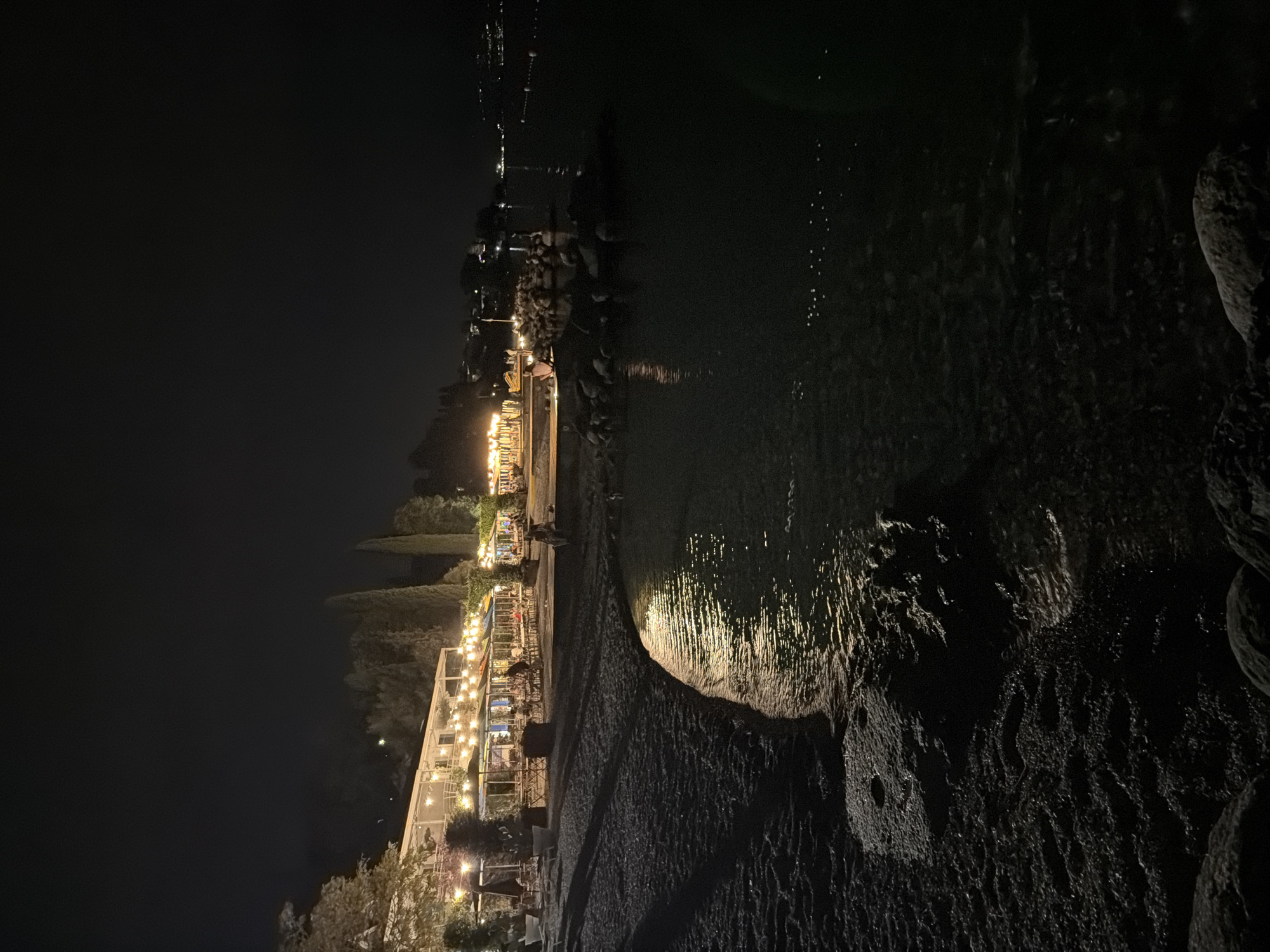 This nighttime photograph captures a scene at a lakeside establishment in Padenghe sul Garda, Italy. The foreground features a pebble beach with the dark, calm water of the lake lapping at its edge, reflecting the artificial lights from the shore. On the left side of the image, a building, likely a restaurant or hotel, is brightly illuminated with strings of lights overhead. Several patrons are visible sitting at tables under umbrellas, indicating outdoor dining. Tall, slender cypress trees are interspersed with the building, adding to the Mediterranean atmosphere. The background shows a darker expanse of the lake stretching towards a distant, dimly lit coastline, suggesting a town or other settlement across the water. A person is seated on a rock structure near the water's edge on the right side of the frame, looking out towards the lake. The overall ambiance is serene and inviting, illuminated by the warm glow of the lights against the dark night sky.