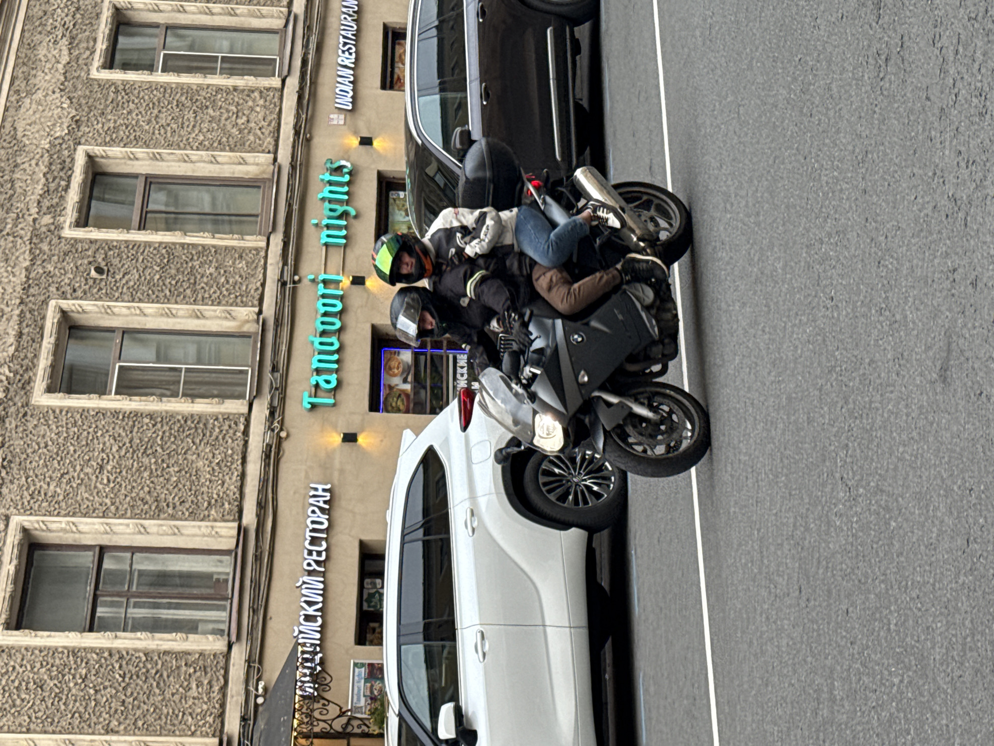 A motorcycle with three people on it is parked on a street in front of a building in Saint Petersburg, Russia. The building has a textured facade and several windows. There are neon signs on the building that read "Tandoori nights" in teal and "ИНДИЙСКИЙ РЕСТОРАН" (Indian Restaurant) vertically in white. There is also a sign that says "INDIAN RESTAURANT" in English. The people on the motorcycle are wearing helmets and jackets. The rider in the front is wearing a green and yellow helmet, and the two passengers behind are wearing dark helmets. One passenger is leaning forward, and the other is leaning back. A white SUV is parked next to the motorcycle. The ground is asphalt with a white line marking the edge of the road. The image appears to have been taken during the day.
