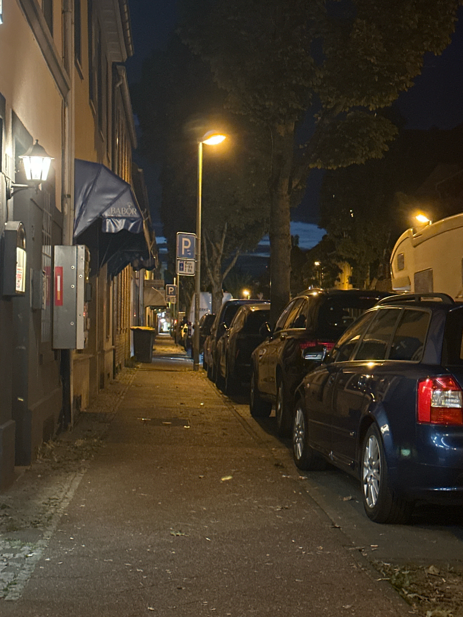 The image depicts a sidewalk and street scene at night in Krefeld, Germany. A row of parked cars lines the right side of the street, with a blue Audi A4 sedan in the foreground. The sidewalk is on the left, with a building facade and a blue awning that reads "BABOR". A streetlamp illuminates the scene. Further down the sidewalk, a dark trash bin is visible. The overall environment suggests a quiet urban street at night.