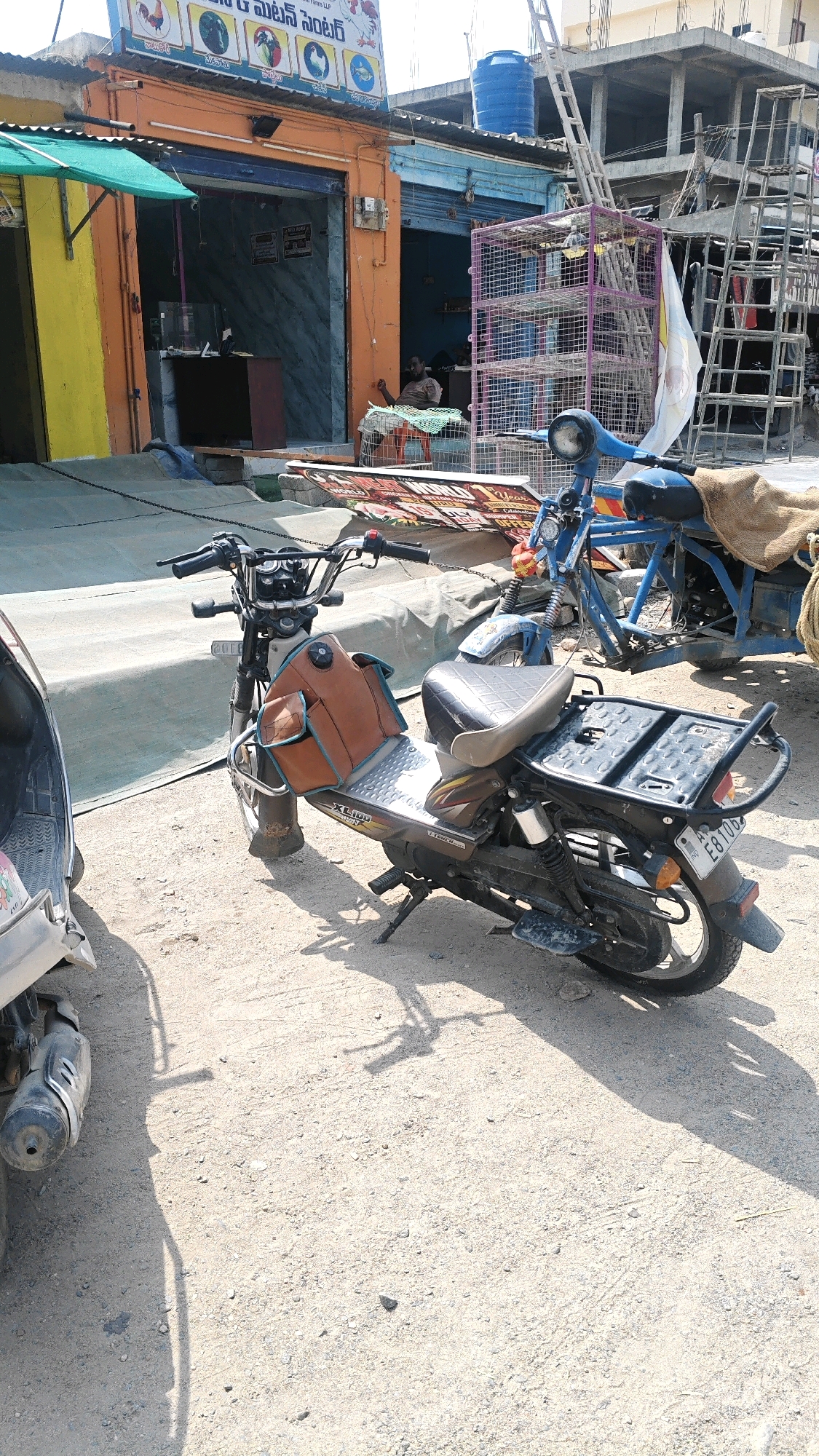 In Pamur, India, a street scene unfolds with a focus on a parked motorcycle. The motorcycle, a dark color with brown saddlebags and a black seat, is positioned on a dirt surface. It features a metal rack at the back. To the left, the front of a silver scooter is visible, casting a shadow. In the background, a building with an orange facade stands, adorned with a sign depicting various birds and fish. Adjacent to this building, a blue water tank is mounted on top of a structure. Further back, scaffolding and partially constructed buildings indicate ongoing development. A man is seated in front of the orange building, partially obscured by a green mat. A purple metal cage stands to the right of the building. The overall environment suggests a commercial area with elements of construction and everyday life.